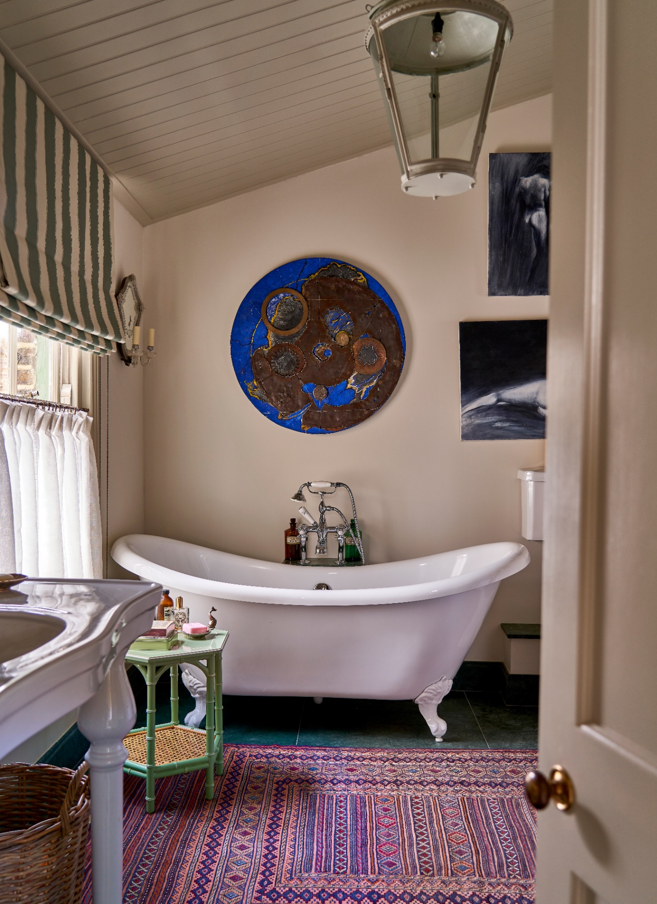 a bathroom with a freestanding bath tub, a large circle piece of art above the bath, a pink and purple patterned rug, a green side table and a window with a green and cream stripe blind