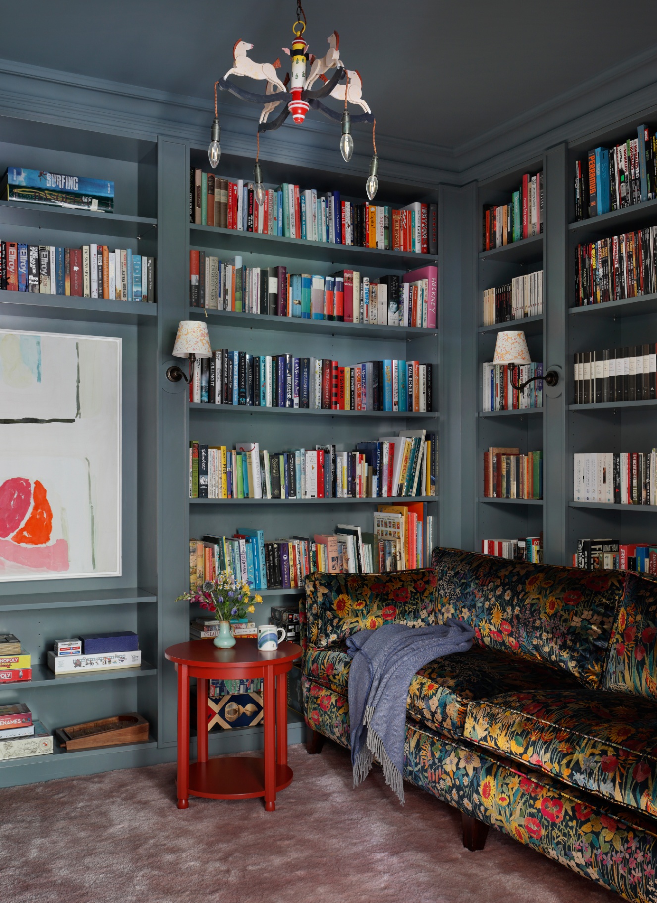 A moody library nook with deep slate-blue painted walls and built-in shelving, featuring a floral velvet sofa, a red side table, and wall sconces with pleated shades
