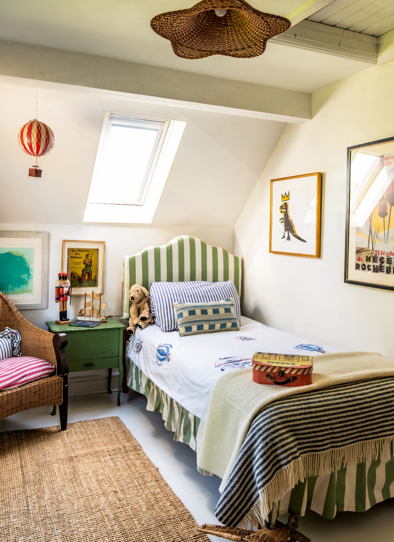 a children's bedroom with a rattan scalloped pendant shade, a red striped hot air balloon hanging from the ceiling and a bed with a grena nd white stripe headboard and valance