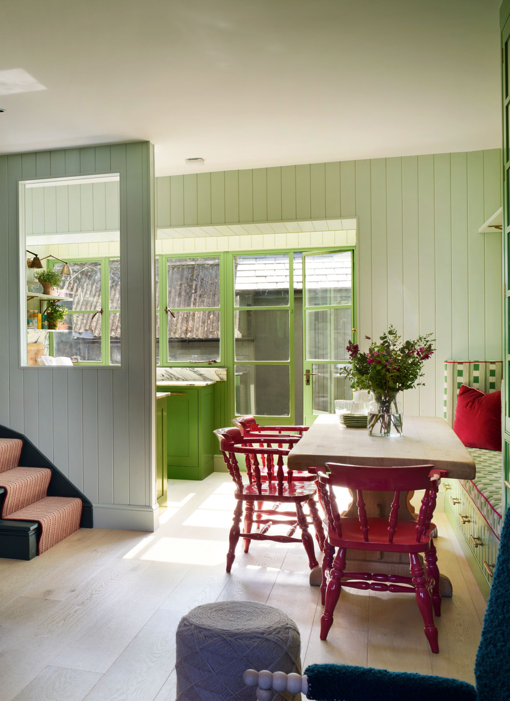 an open plan dining room and kitchen with awooden dining table and bright red dining chairs and sage green panelled walls