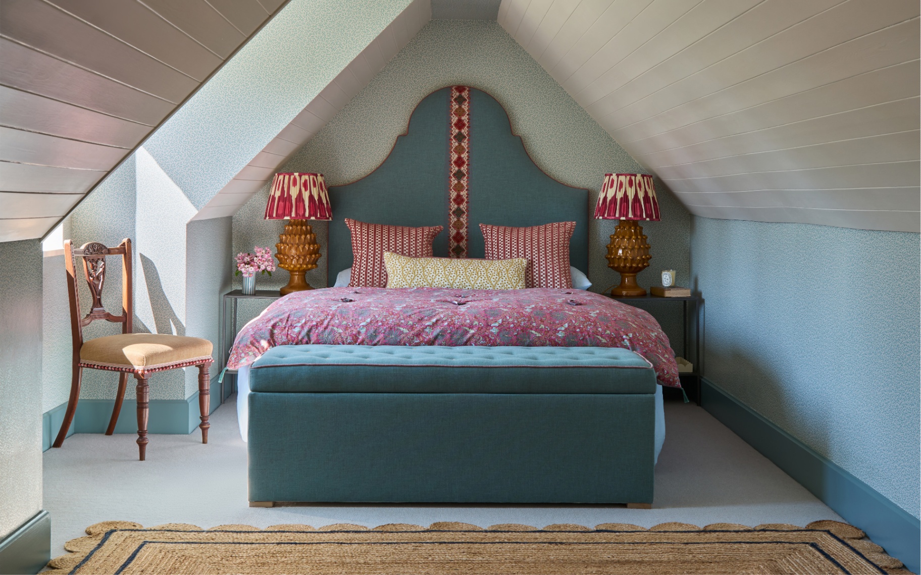 an attic bedroom with white paneled ceilings and blue wallpaper on the walls, a large bed in the middle with a blue and red patterned headboard, red and yellow cushions, a pink and blue floral bedspread, a large blue ottoman at the foot of the bed