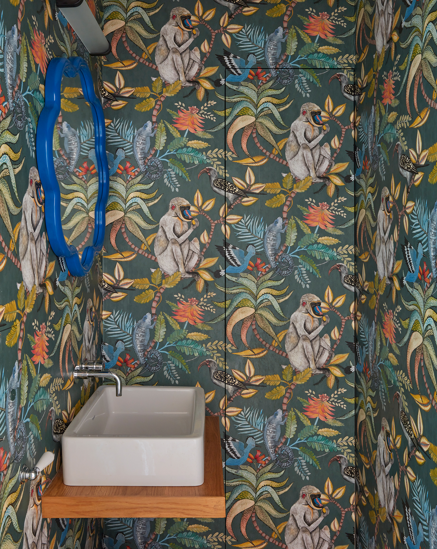 A small cloakroom with a colourful jungle-themed wallpaper, a jib door and a simple basin on a floating wooden shelf. Above hangs an electric blue flower-shaped mirror and contemporary wall light