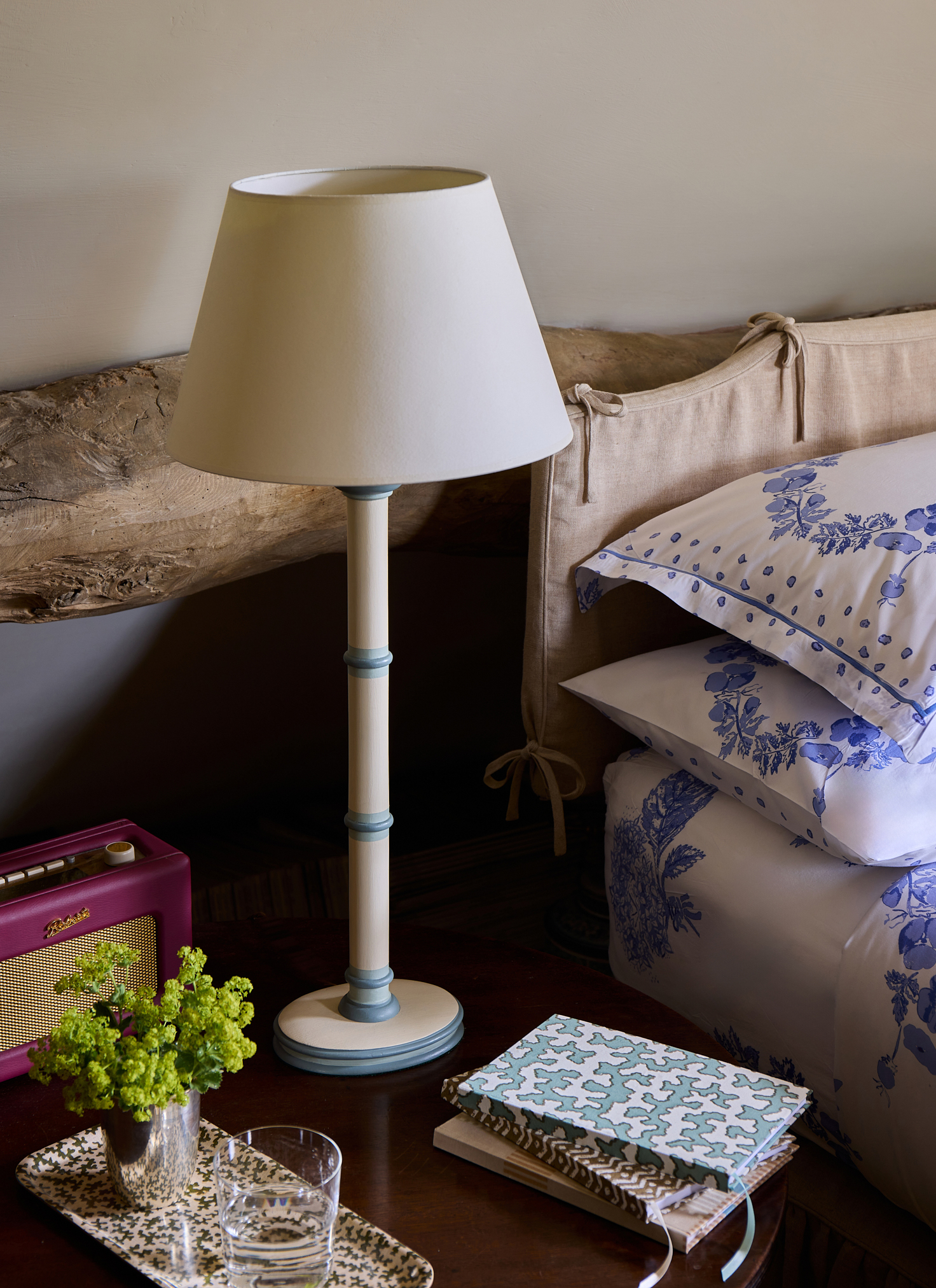 close up of a bedside table with a cream lamp, a dark red roberts radio, a pile of notebooks and a tray with a glass of water
