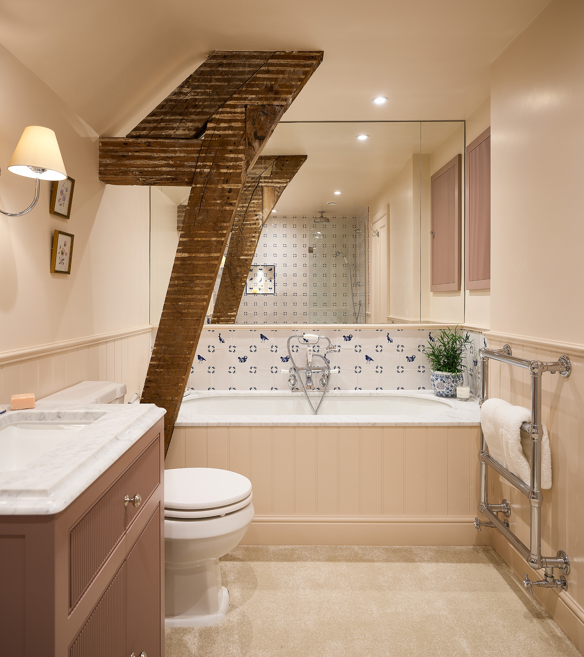 a loft bathroom with wooden beams, a bath tub with a panel wall mirror above, a silver heated towel rail and a mauve vanity unit with a white sink