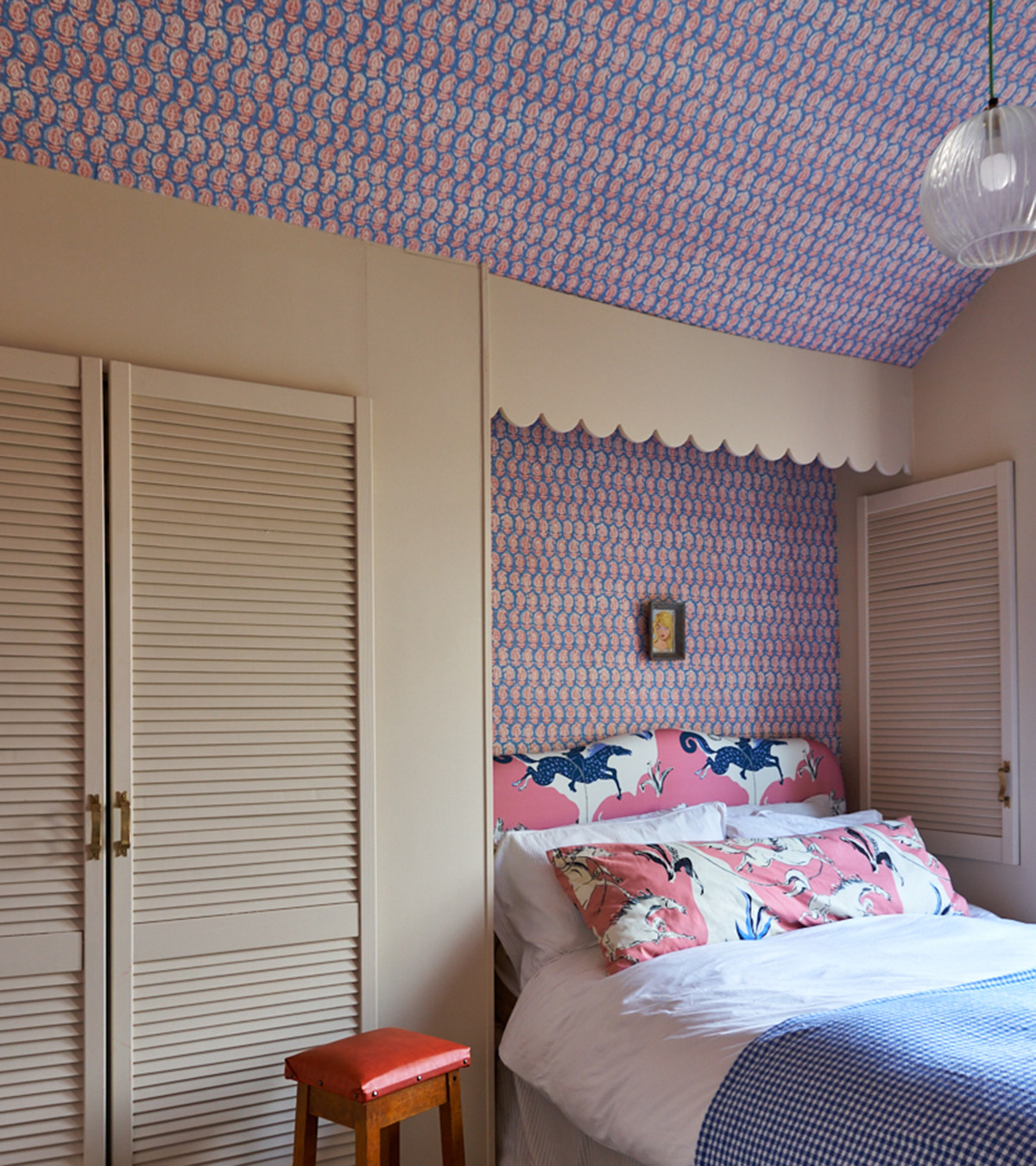 a bedroom with purple floral wallpaper behind the bed and on the ceiling, cream built in cupboards and a single bed with white bedding, a blue bed spread and pink headboard