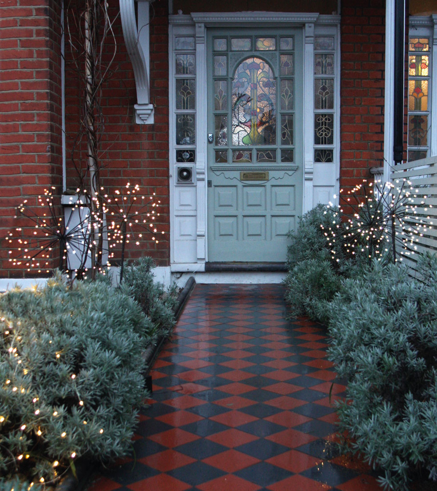 a front of a house with a sage green door, a red and black checkerboard path leading up to the house with bushes lining it with led solar stake lights