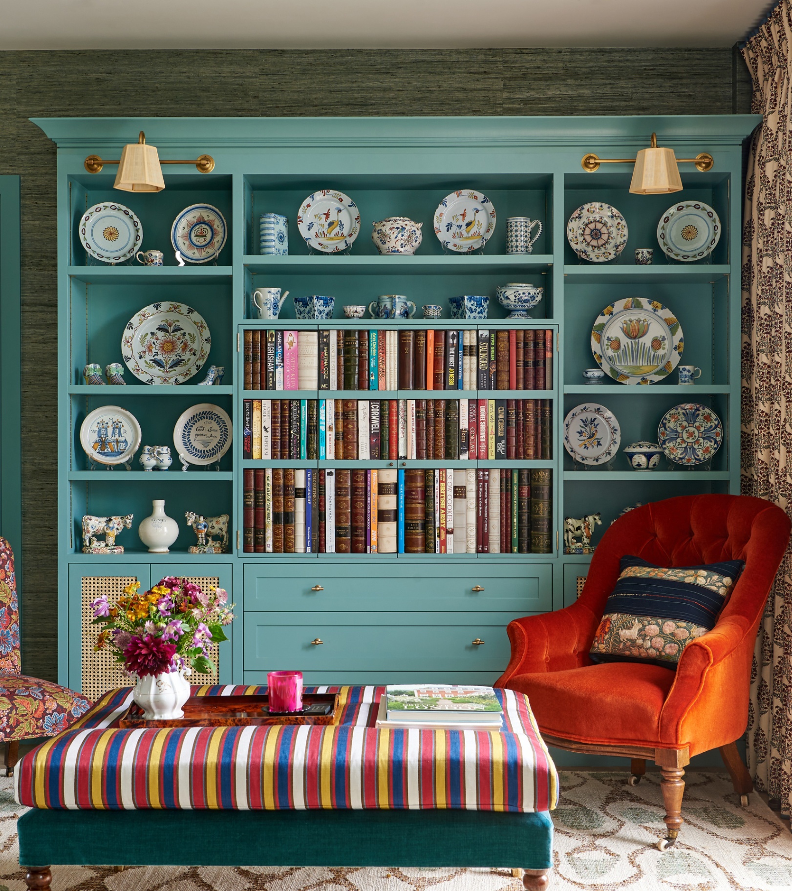 a living room with a large pale blue painted bookshelf with vintage books in the middle and patterned china all around the outside shelves, a multicoloured striped ottoman and an orange velvet armchair next to it
