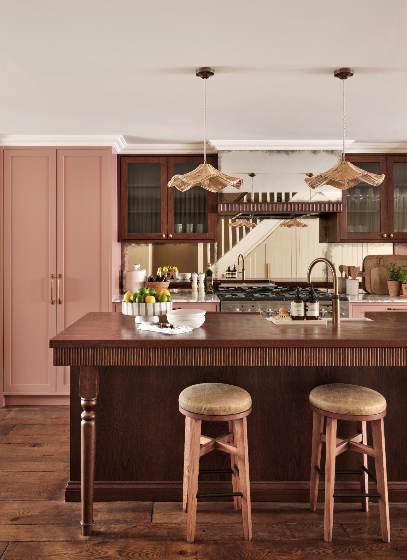 a kitchen with wooden floors, dark wooden kitchen island and floating cupboards, rattan pendants above the island and a cupboard painted in pale pink