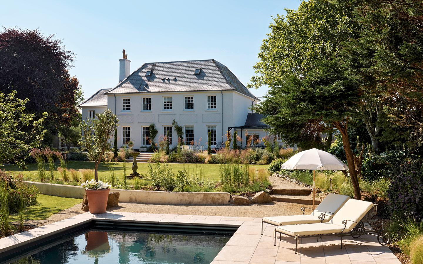 A large georgian home sits in the background with a manicured lawn and landscaped garden in front. In the foreground the end of a swimming pool can be seen, along with two sun loungers under a parasol