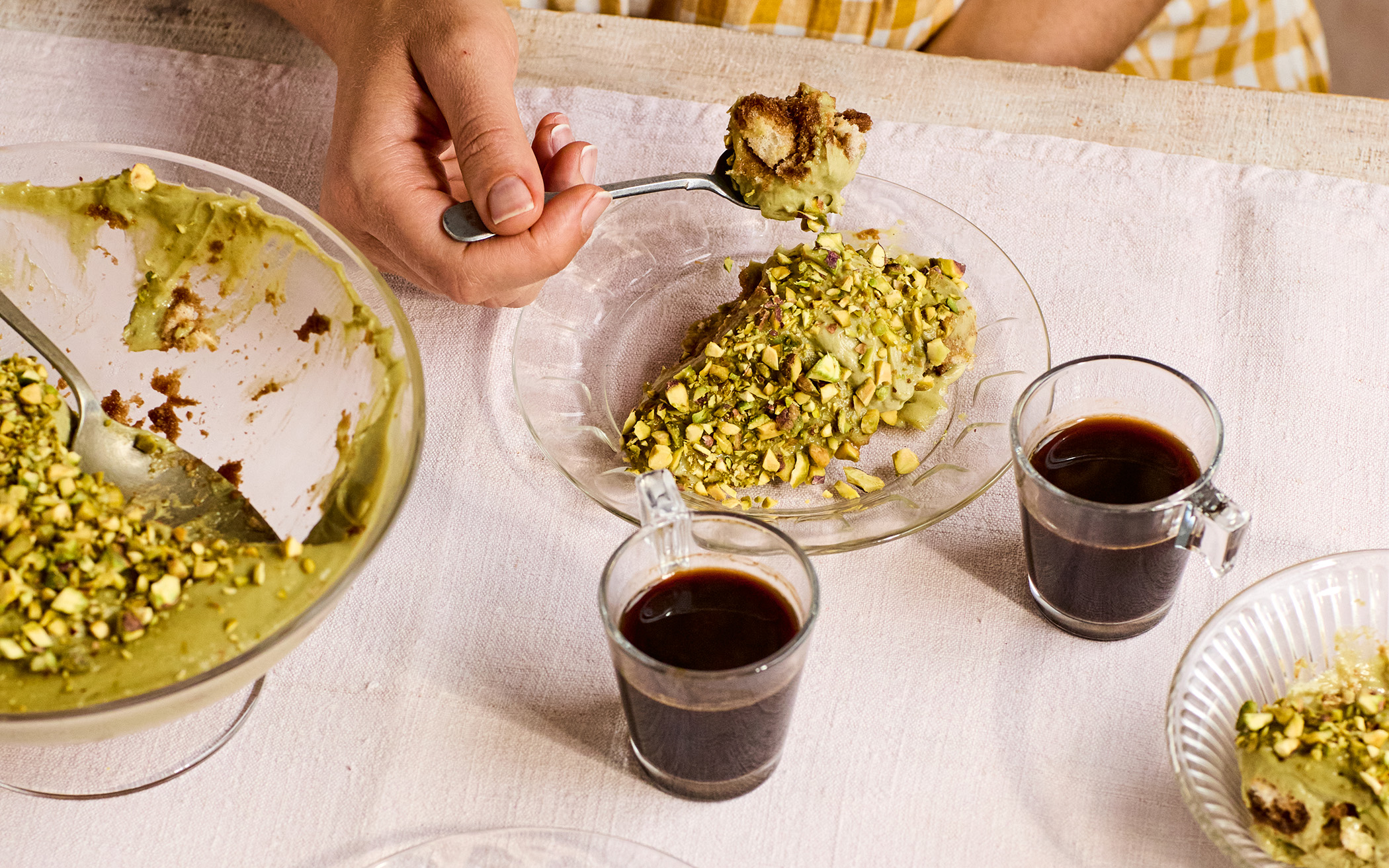 pistachio tiramisu in a serving bowl on a white table cloth with two cups of black coffee