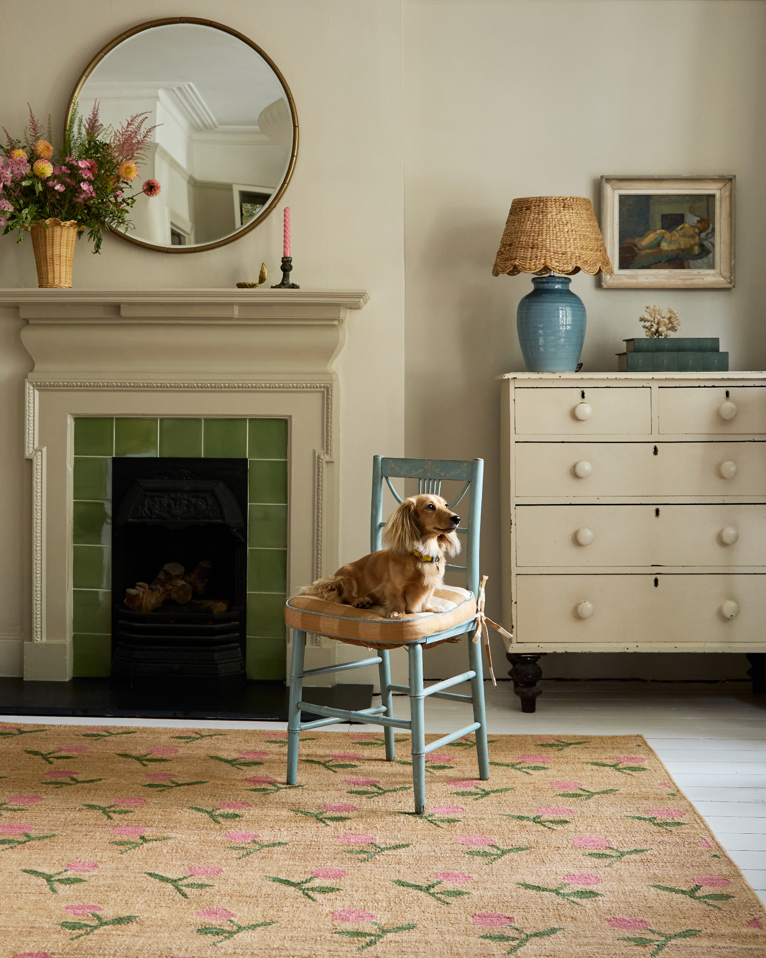a living room with a green tiled fireplace, a circle mirror adn a blue dining chair with a long haired dachsund sat on it
