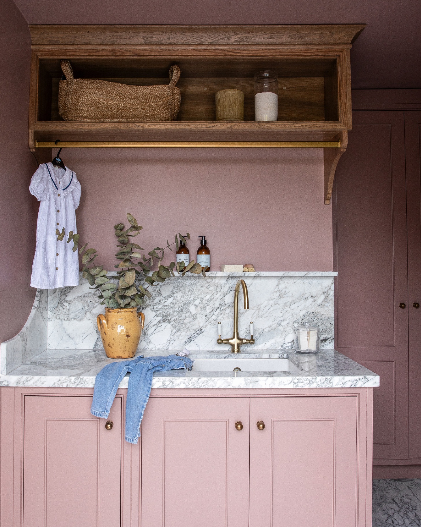 a utility room with pale pink walls and cabinets, a white marble countertop and splash back and a large wooden open shelf near the ceiling