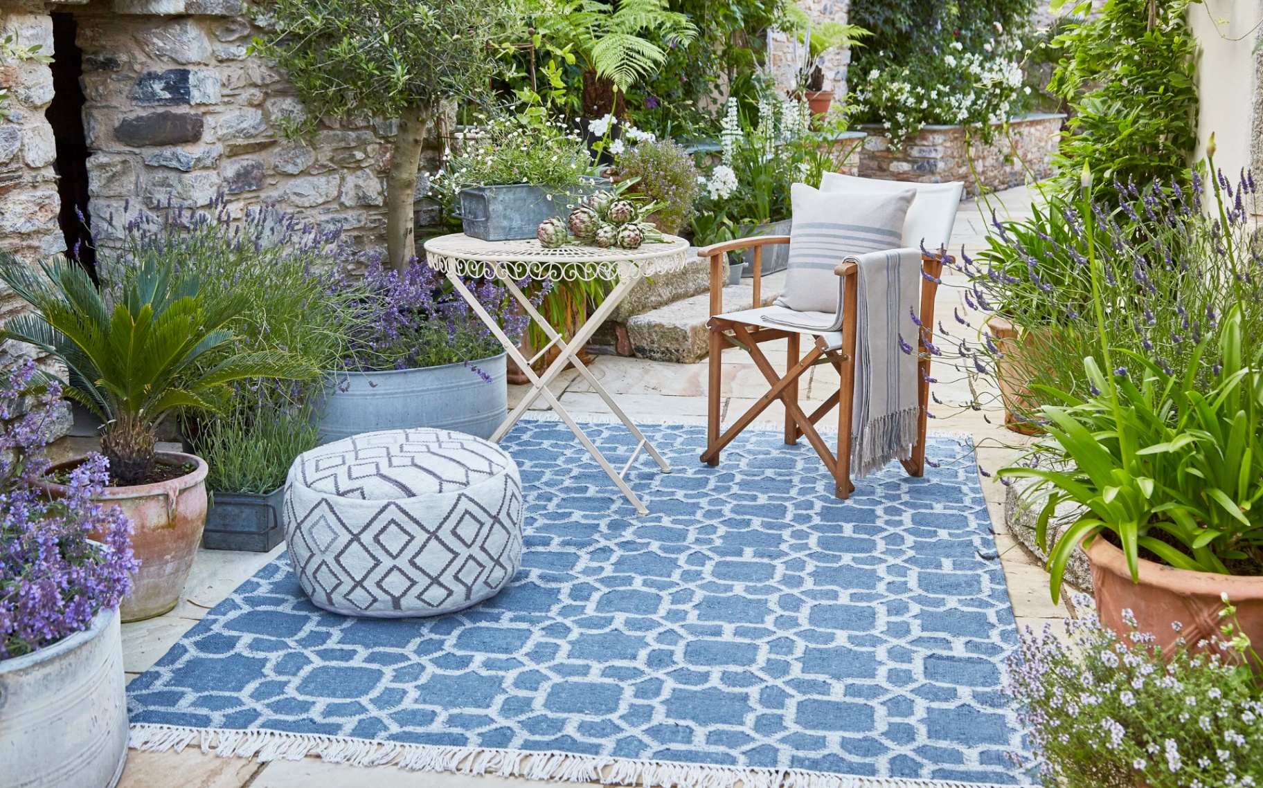 a blue patterned outdoor rug on the floor of a courtyard garden, with  a cream deckchair with outdoor cushions surrounded by plant pots filled with greenery