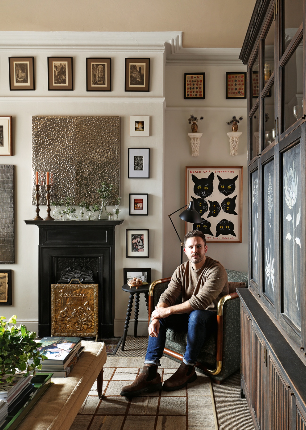 Interior designer Christian Bense sits on a midcentury armchair in his living room in front of walls decorated with an eclectic mix of artwork and beside a fireplace with an ornate brass fire screen