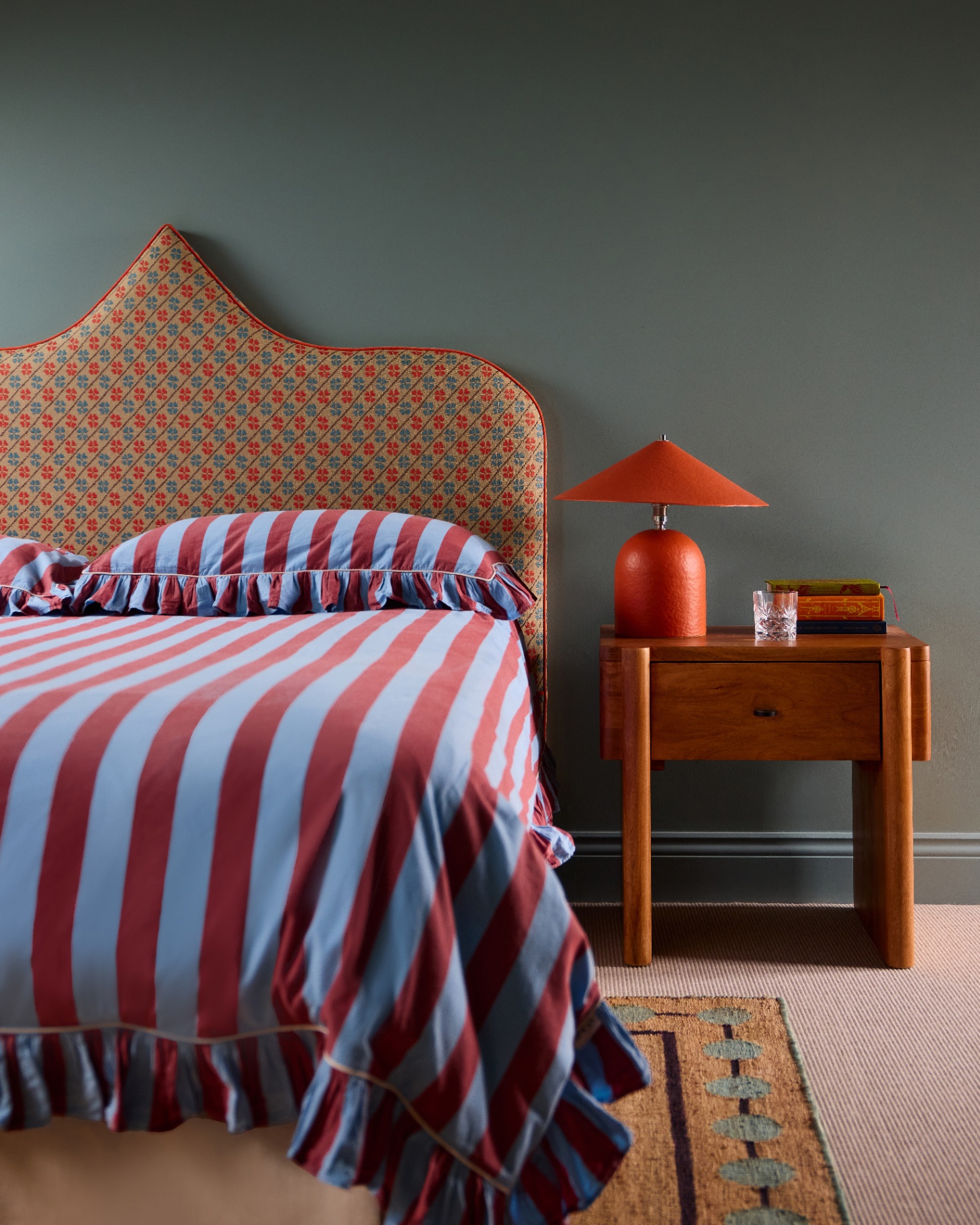 a bedroom with moss green walls, a bed with a an orange patterned headboard and dark red and blue striped ruffle bedding and a large wooden bedside table