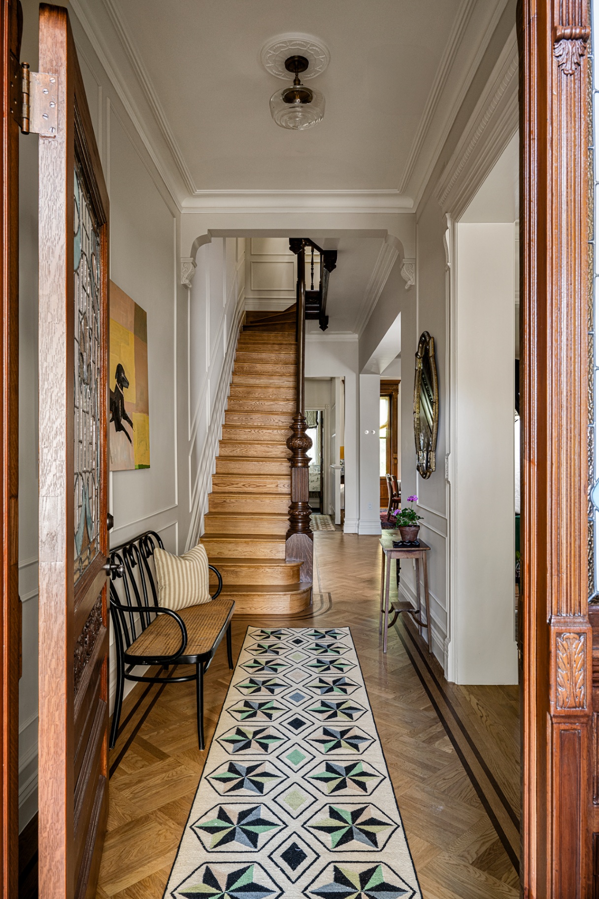Looking through a dark wood front door to an entrance hall with a geometric star runner on the herringbone wood floor. A bench sits to the left while wooden stairs lead up to the next floor in the background