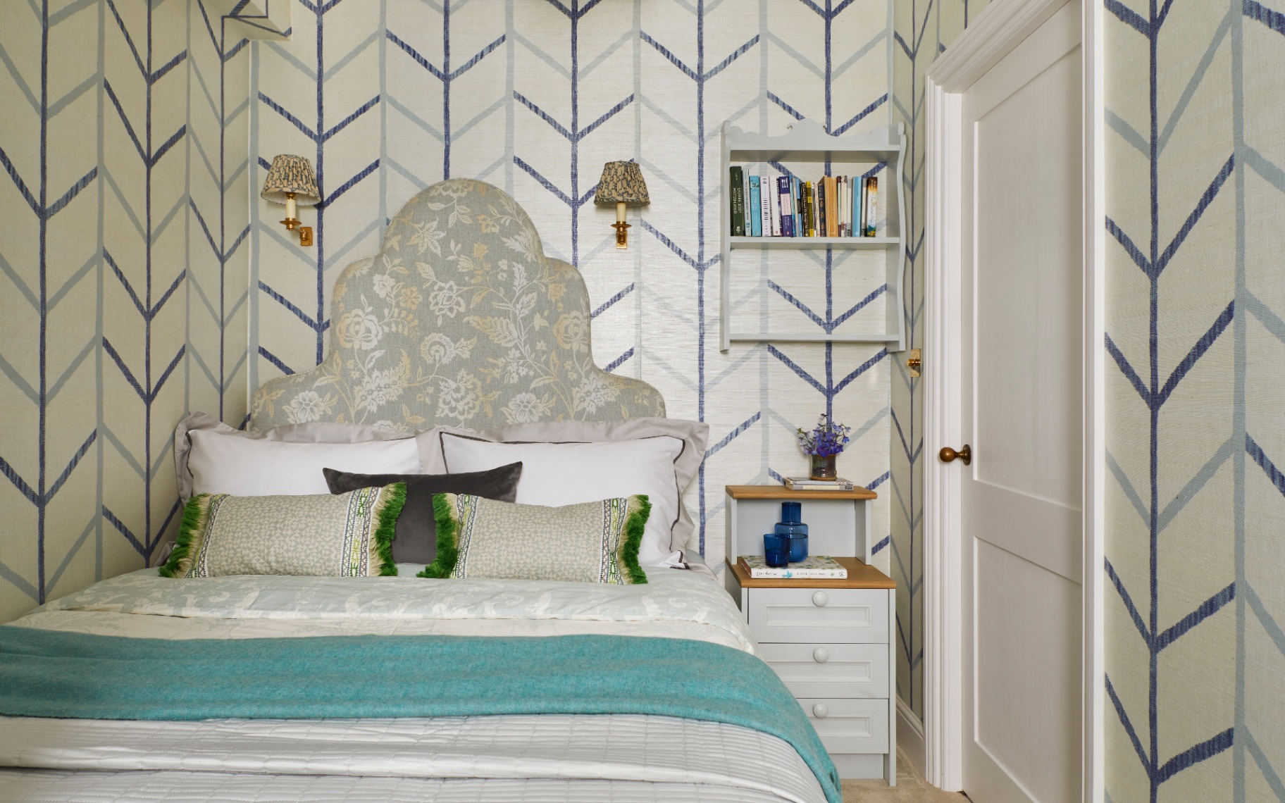 A SMALL guest bedroom with white and blue stripe chevron wallpaper, a bed with a large sage green patterned headboard and white bedding and a small white shelving unit