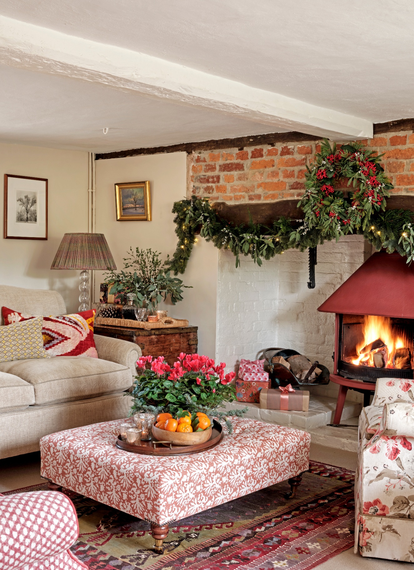 A cozy living room with a brick fireplace, a burning fire, and garlands on the mantel, featuring a patterned ottoman with a tray holding a bowl of vibrant pink potted cyclamen