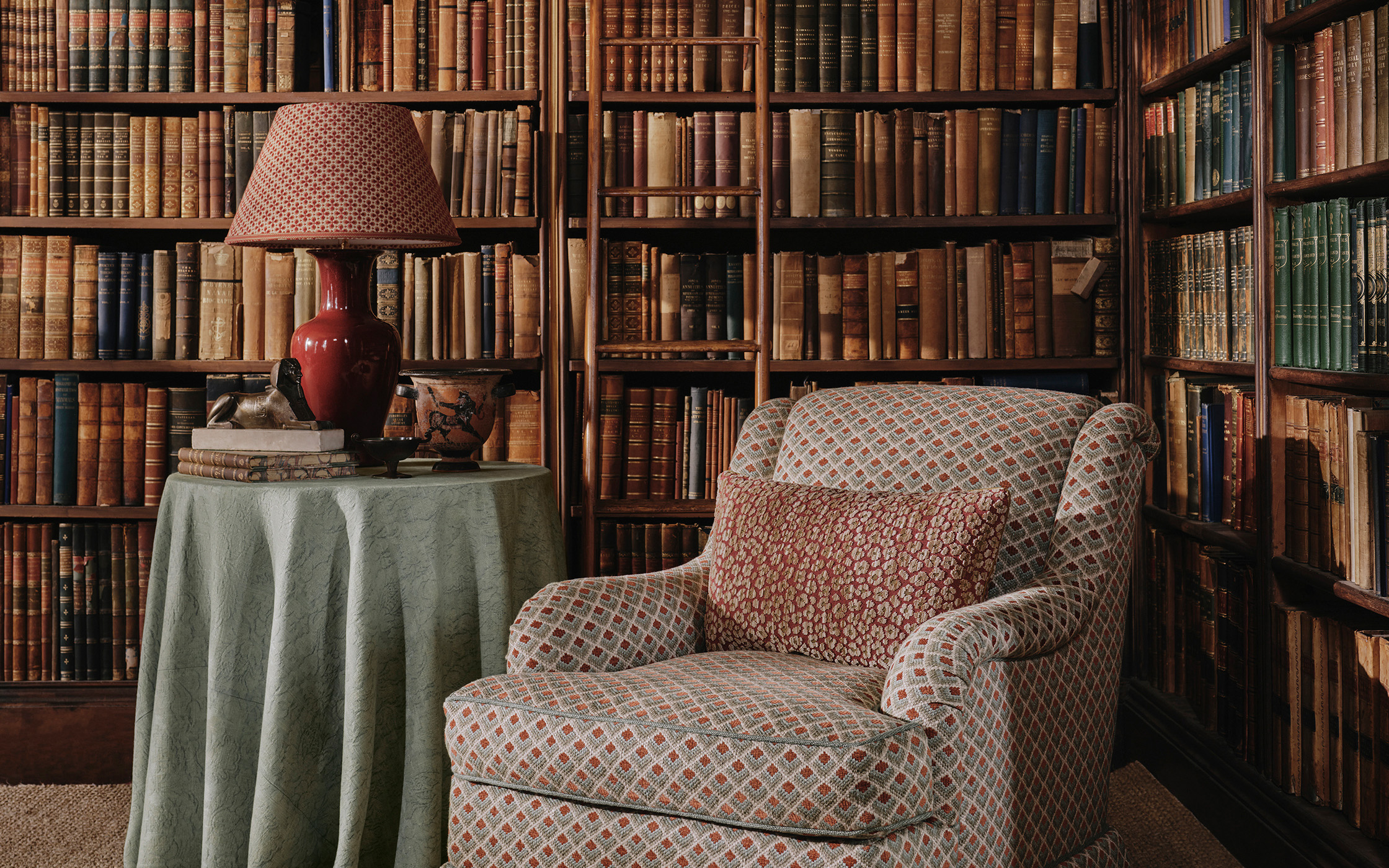 a patterned arm chair with a side table next to it draped in a pale blue cloth with a large wraparound brown book shelf behind filled with antique books