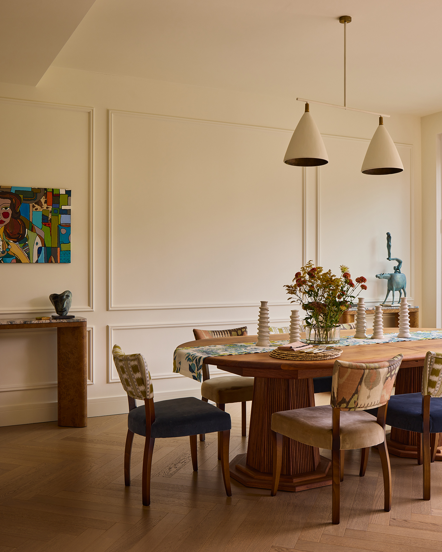 A dining room with a large oval wooden table and upholstered dining chairs. The table is topped with vases, candlesticks, a runner and a stack of placemats, napkins and cutlery. A two-light pendant hangs above. Panelled walls are painted cream
