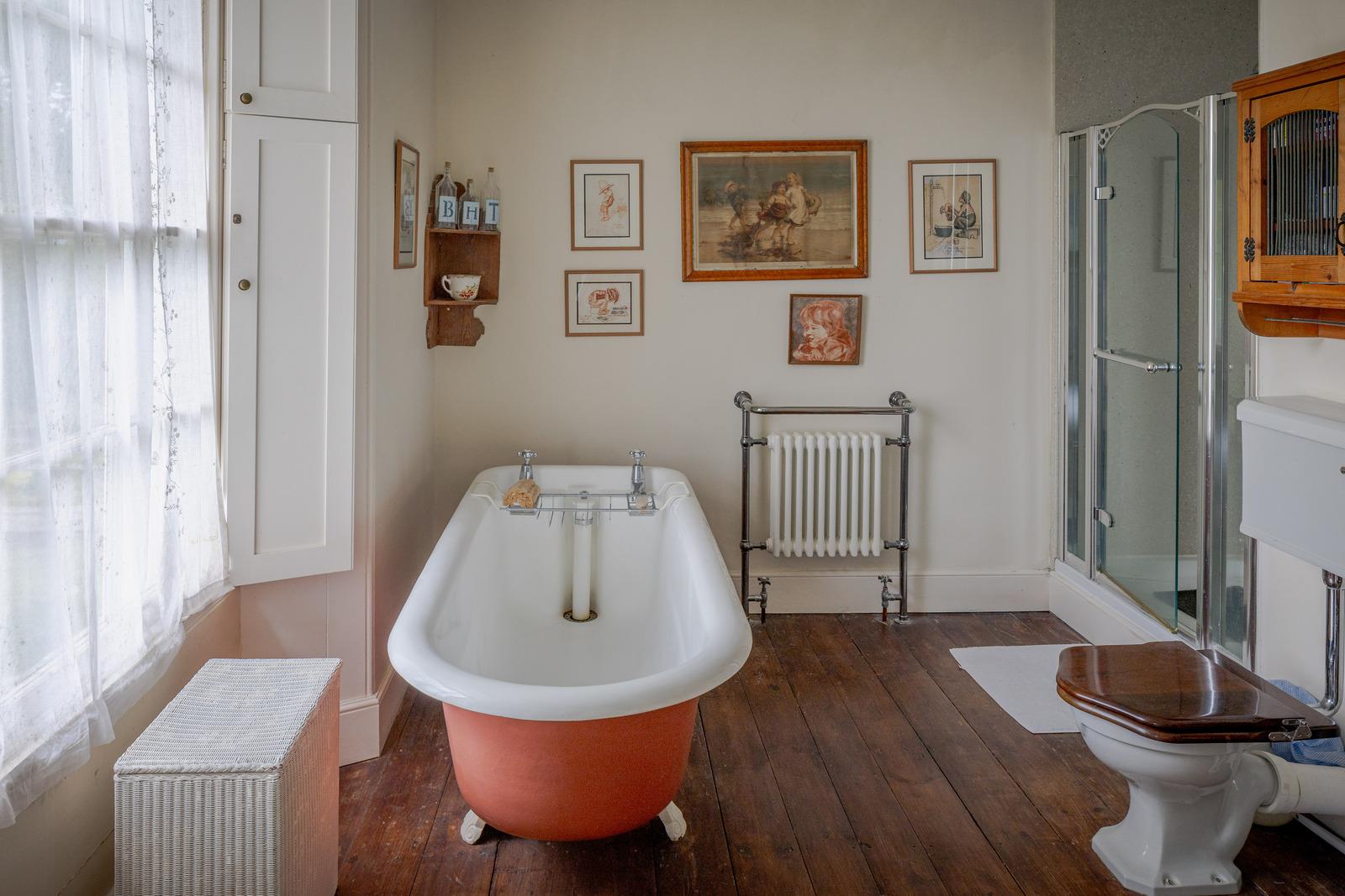 a bathroom with a coral pink freestanding bath, wooden floors, white walls and a collection of paintings hung on the wall