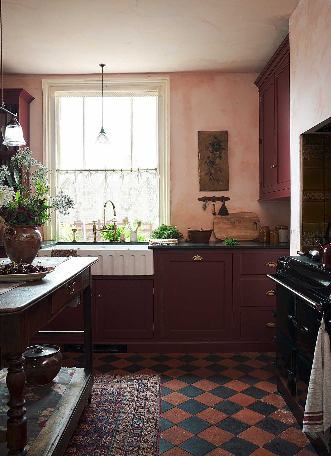 a kitchen with burgundy cabinets, black and terracotta flooring, a kitchen island and a belfast sink in front of a window