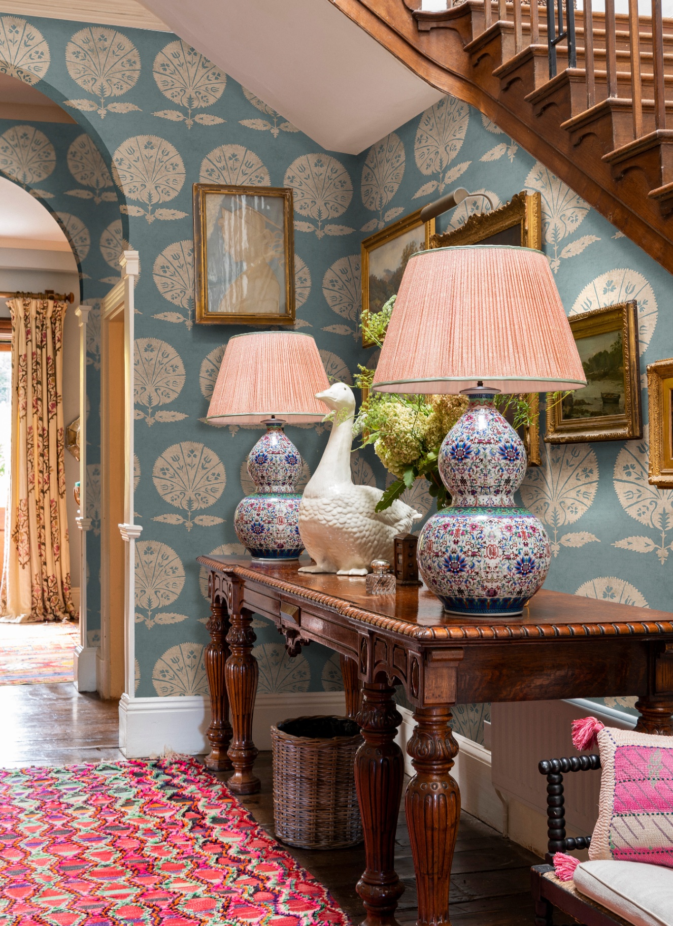 a hallway with blue and white floral wallpaper, a red patterned runner rug and a large antique dark wooden console table with two large lamps and a statue of a duck