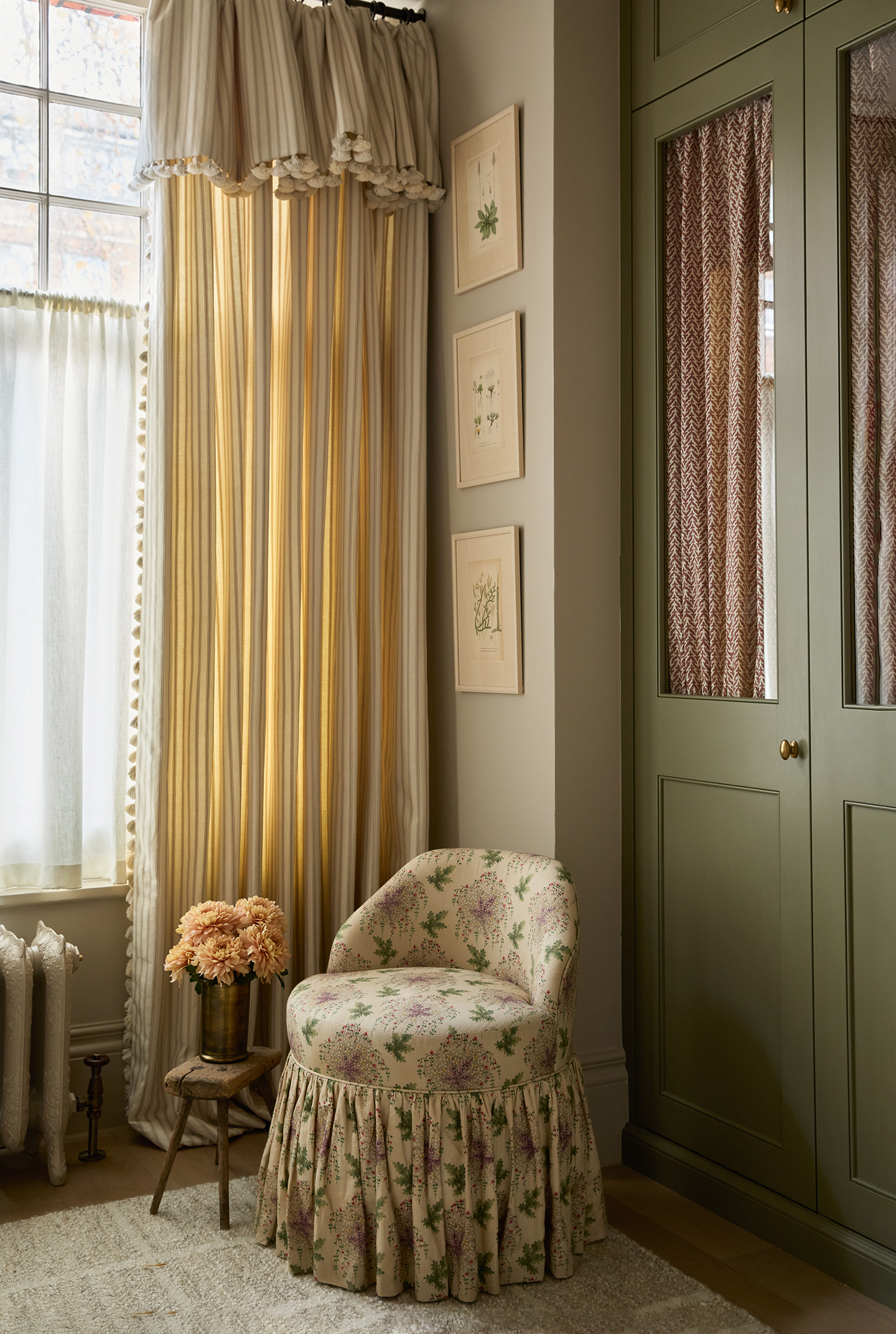 The corner of a bedroom with a built in green wardrobe, frilled accent chair and striped curtain with a flop over header