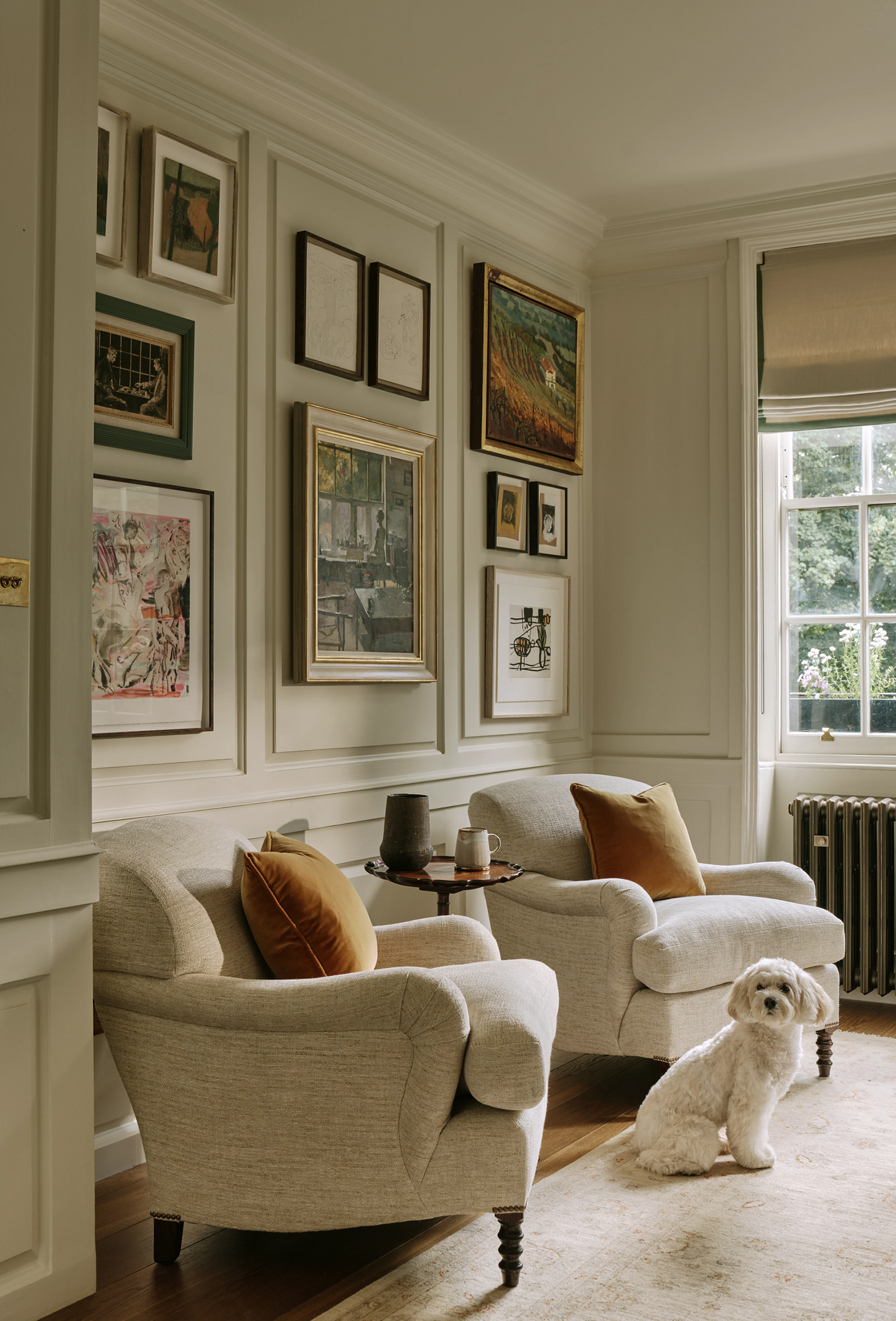 Two neutral armchairs dressed with matching orange velvet pillows, arranged beneath a panelled wall covered in a range of artworks. A white dog sits in front of the chairs on a faded patterned neutral rug
