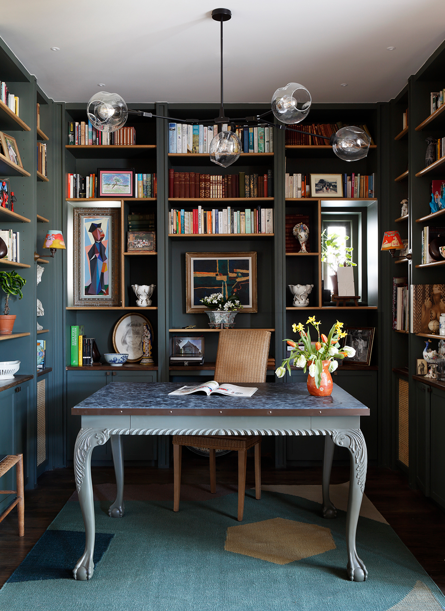 Dark teal green library study with floor-to-ceiling built-in bookcases, a grey-painted desk floating in the center, glass globe chandelier, and blue-green rug
