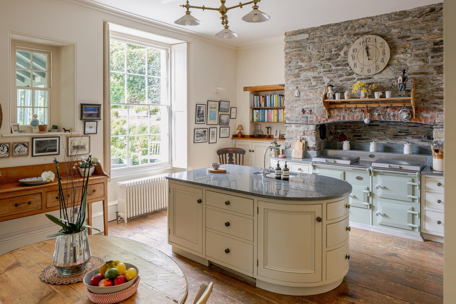 a country kitchen with a sage green range cooker with exposed brick surround, a cream kitchen island and a light fitting with three shades