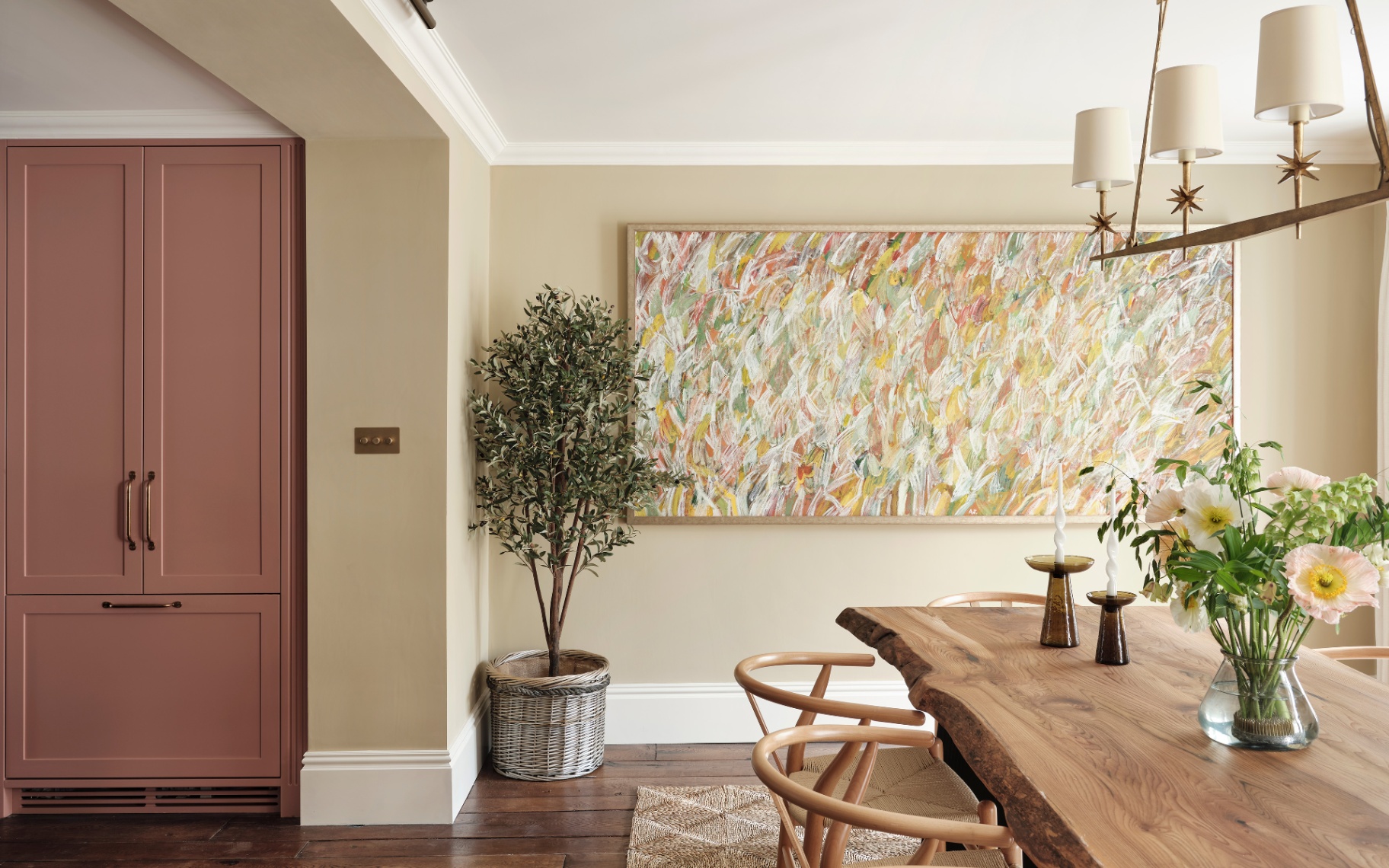 a dining room with dark cream walls, a large abstract painting on the wall with a potte dolive tree on the floor in front it it, a mauve painted cabinet to the left and a large wooden dining table and chairs with flowers on top and a chandelier above