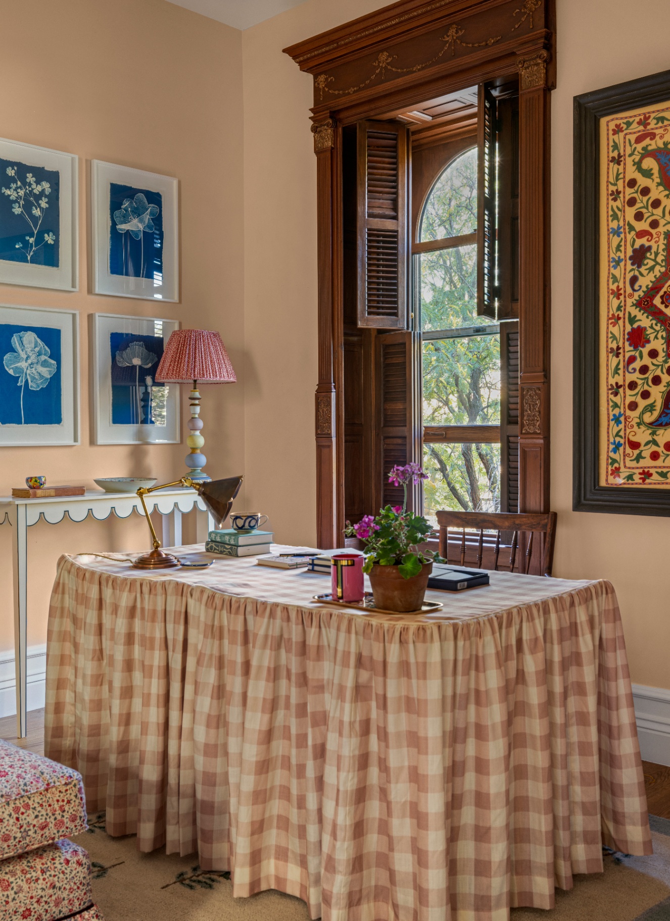 Traditional home office featuring a work table draped in a full-length pink and white ruffled check tablecloth, framed cyanotype art, and tall dark wood window shutters