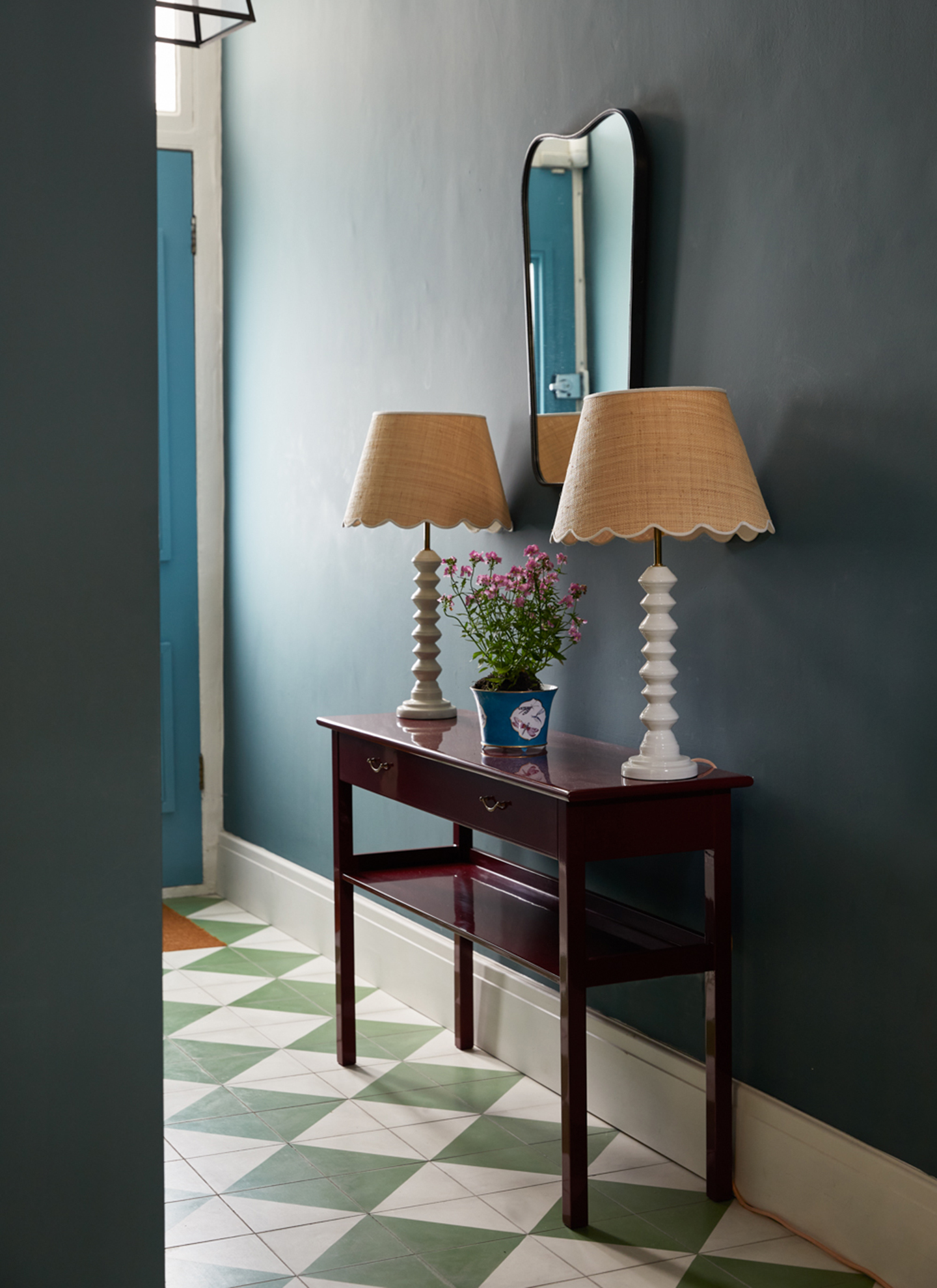 a hallway with dark navy walls, a glossy dark purple console with two matching white adn raffia lamps and a green and white checkerboard floor