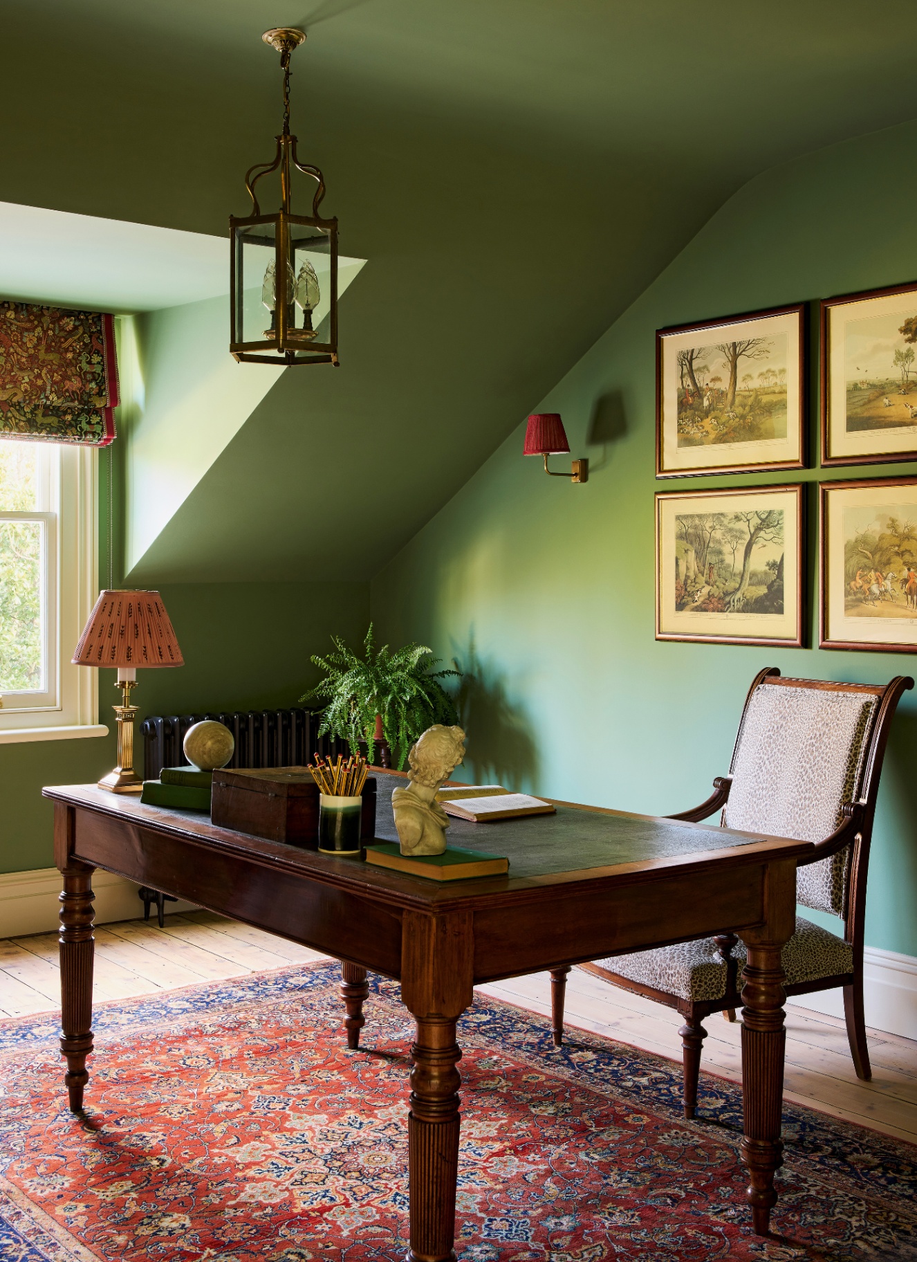 Attic study painted in deep sage green across walls and sloped ceilings, featuring a centered wood executive desk, red Persian rug, and brass lantern light