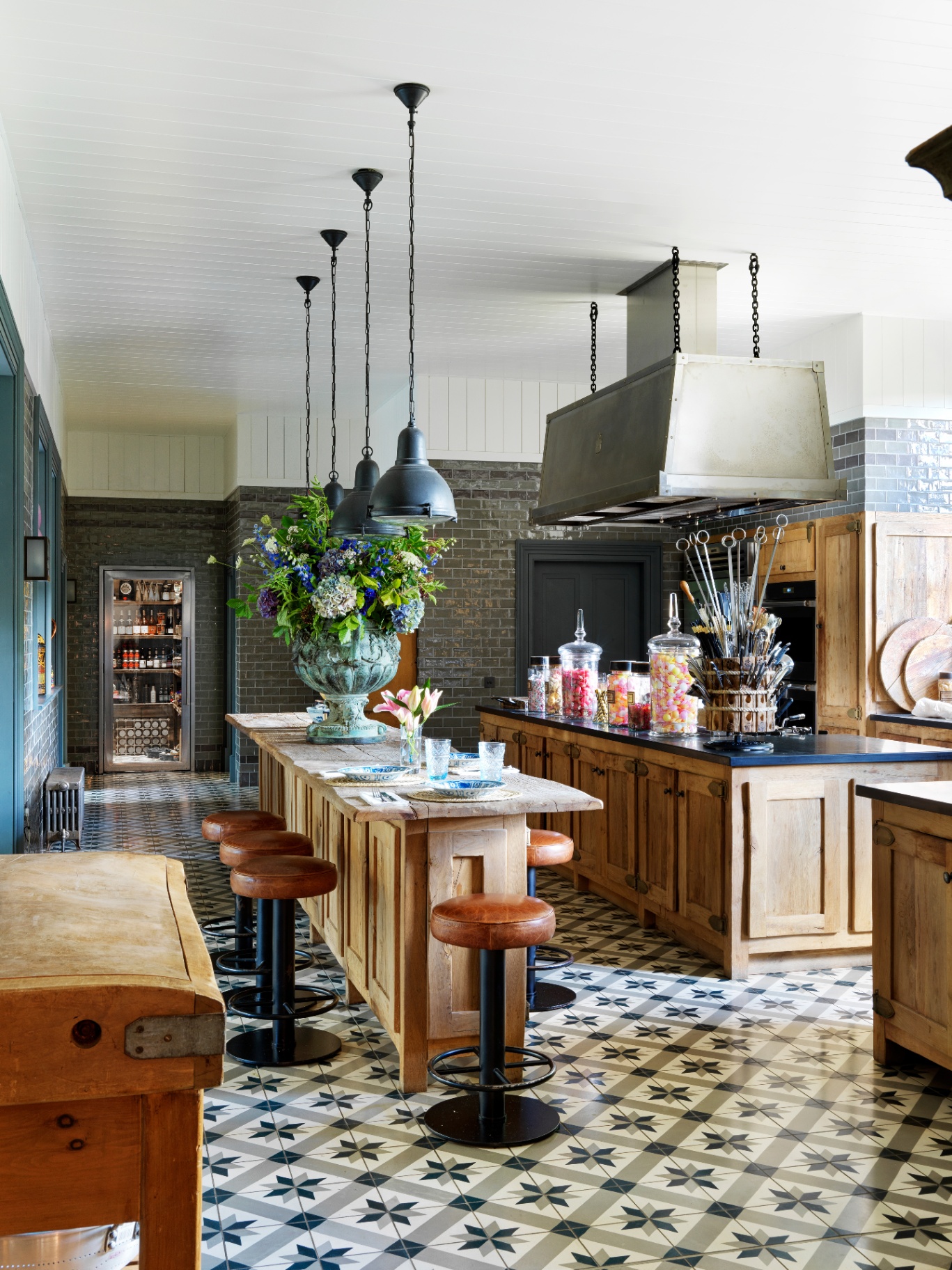 Jade Holland Cooper’s spacious country kitchen with long wooden islands, leather bar stools, tiled flooring, and a large floral centerpiece