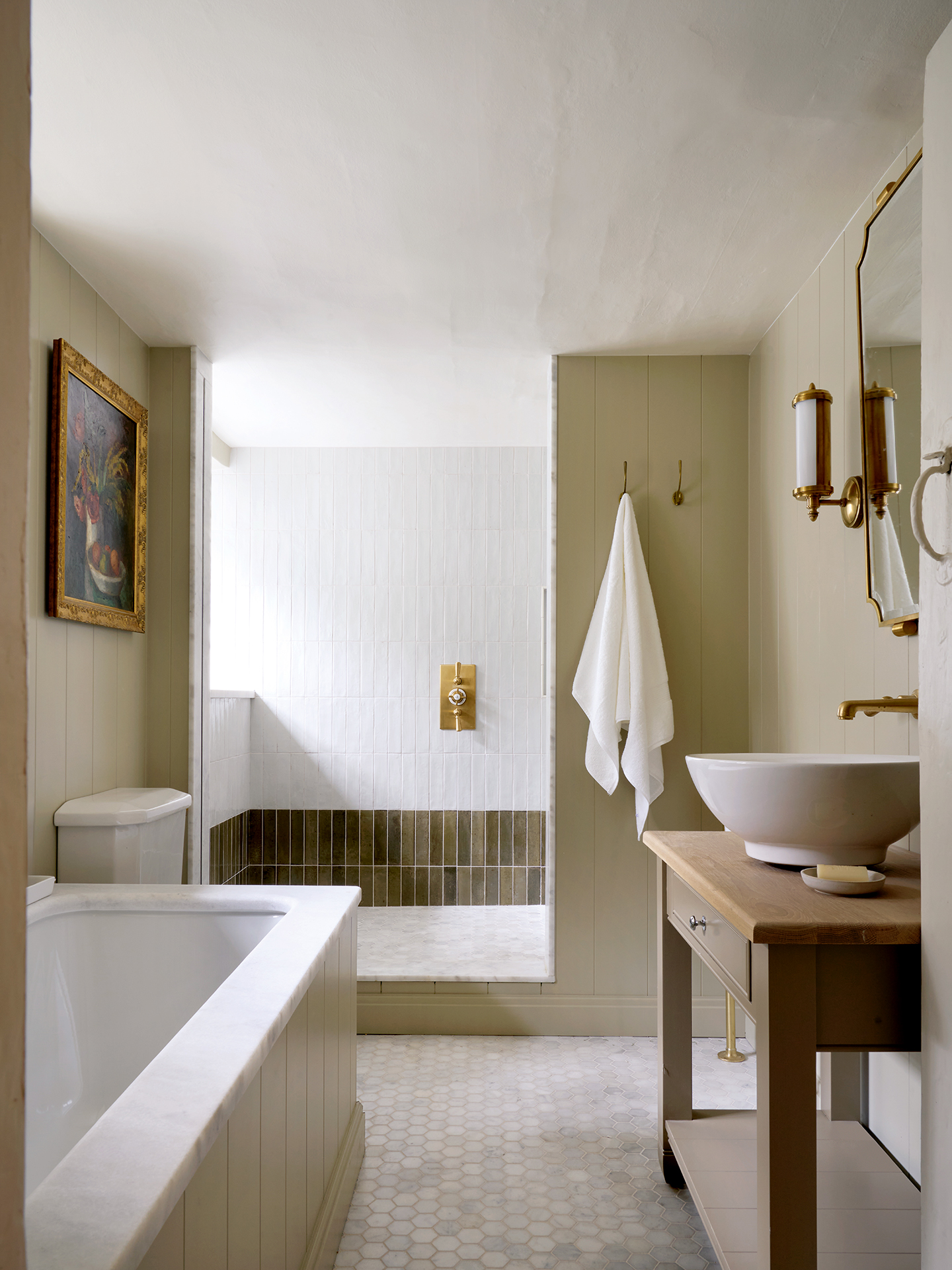 A bathroom with a tongue-and-groove built-in bath to the left and a wooden washstand with a curved basin to the right. In the background is a shower area tiled with white metro tiles on the upper two thirds of the wall and green metro tiles on the lower