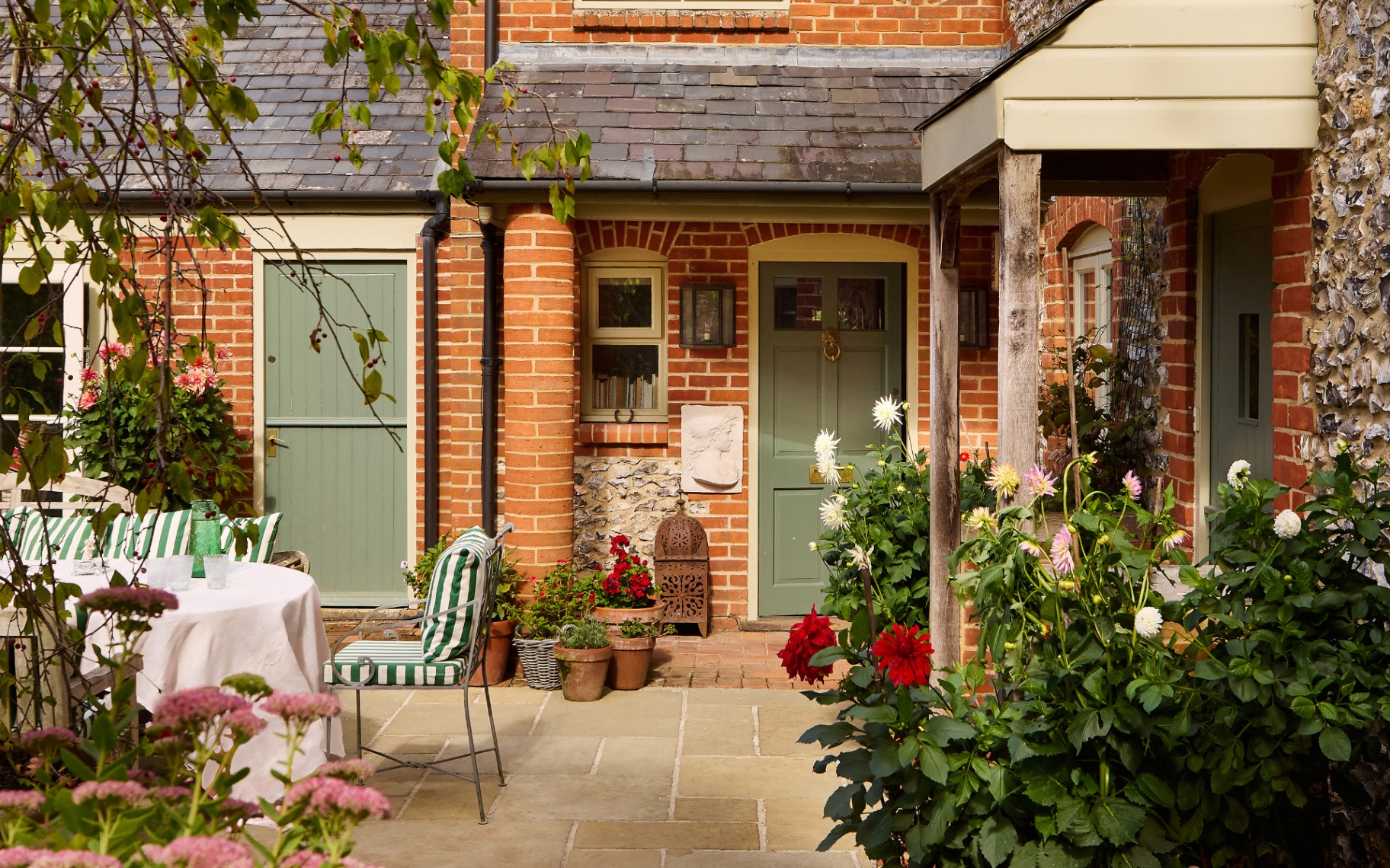 front of a red brick house with a green door, a porch, talll colourufl flowers and an outdoor dining set on a patio with white and green striped cushions