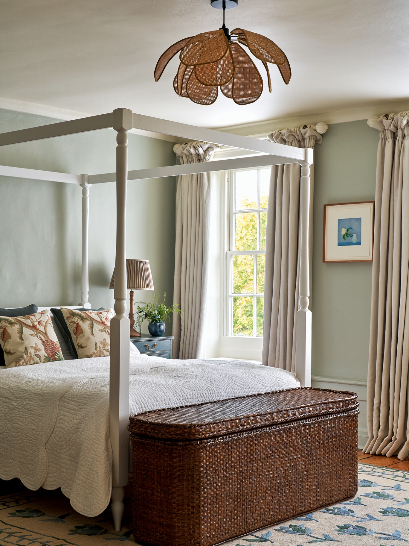 A blue-green painted bedroom with a white four-poster bed, decorated with floral cushions and a white patterned quilt, a rattan storage bench at the end of the bed, a rattan flower-style pendant and neutral pleated curtains 