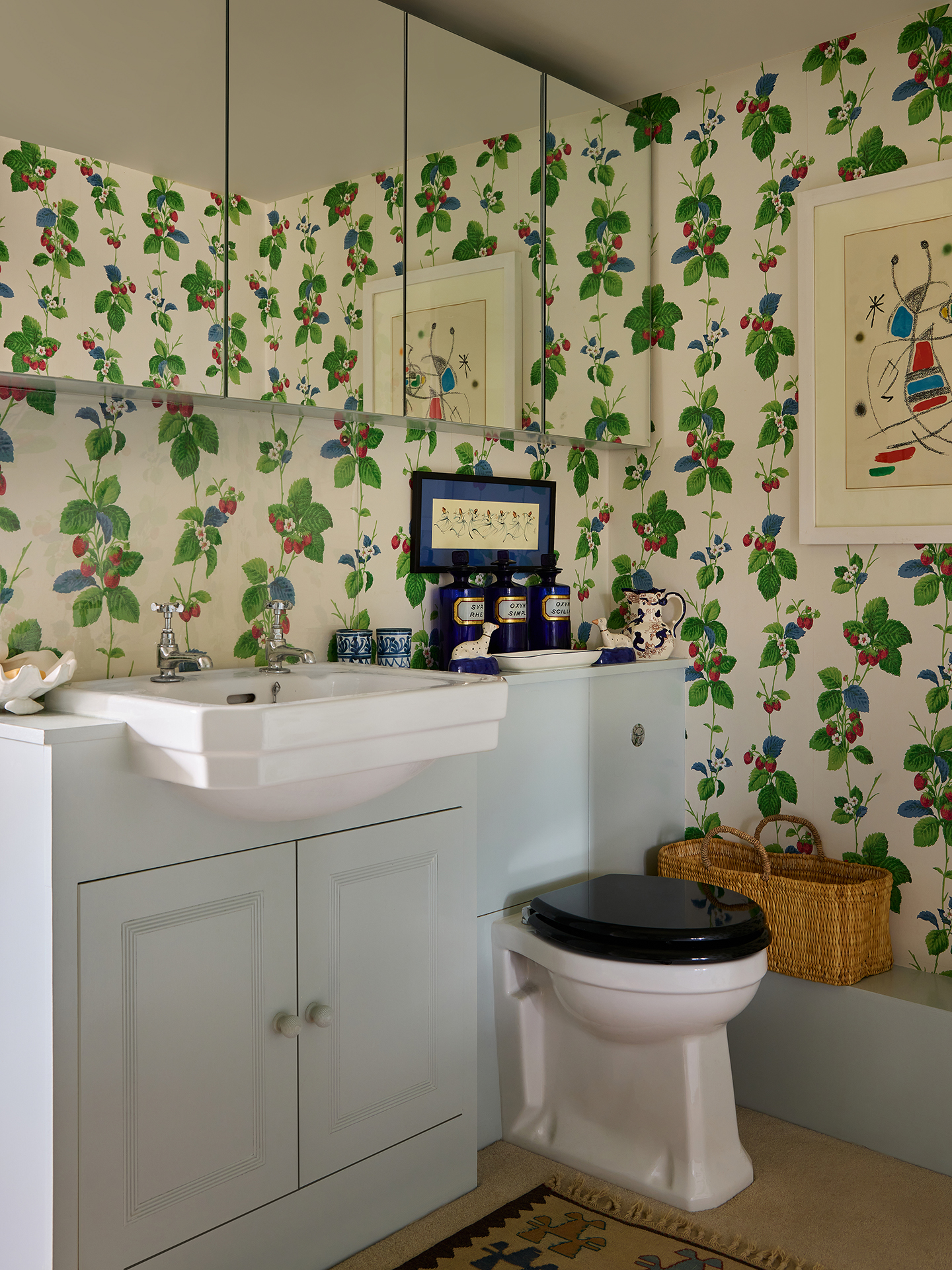 A bathroom with a toilet and built-in vanity unit with basin. Mirrored cabinets line the upper part of the wall and the walls are covered in a strawberry-themed wallpaper