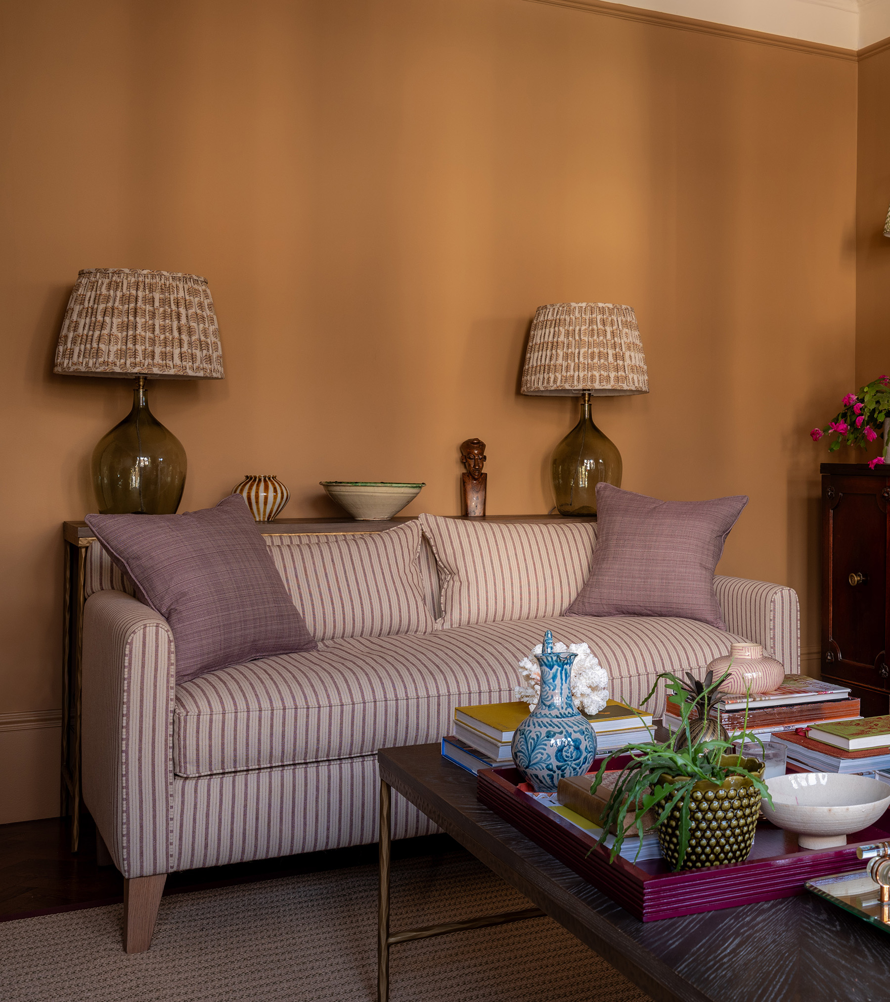 a living room with ochre walls, a white and pink stripe sofa with a console behind it with two matching lamps and a coffee table in front of it with a burgundy tray and a plant