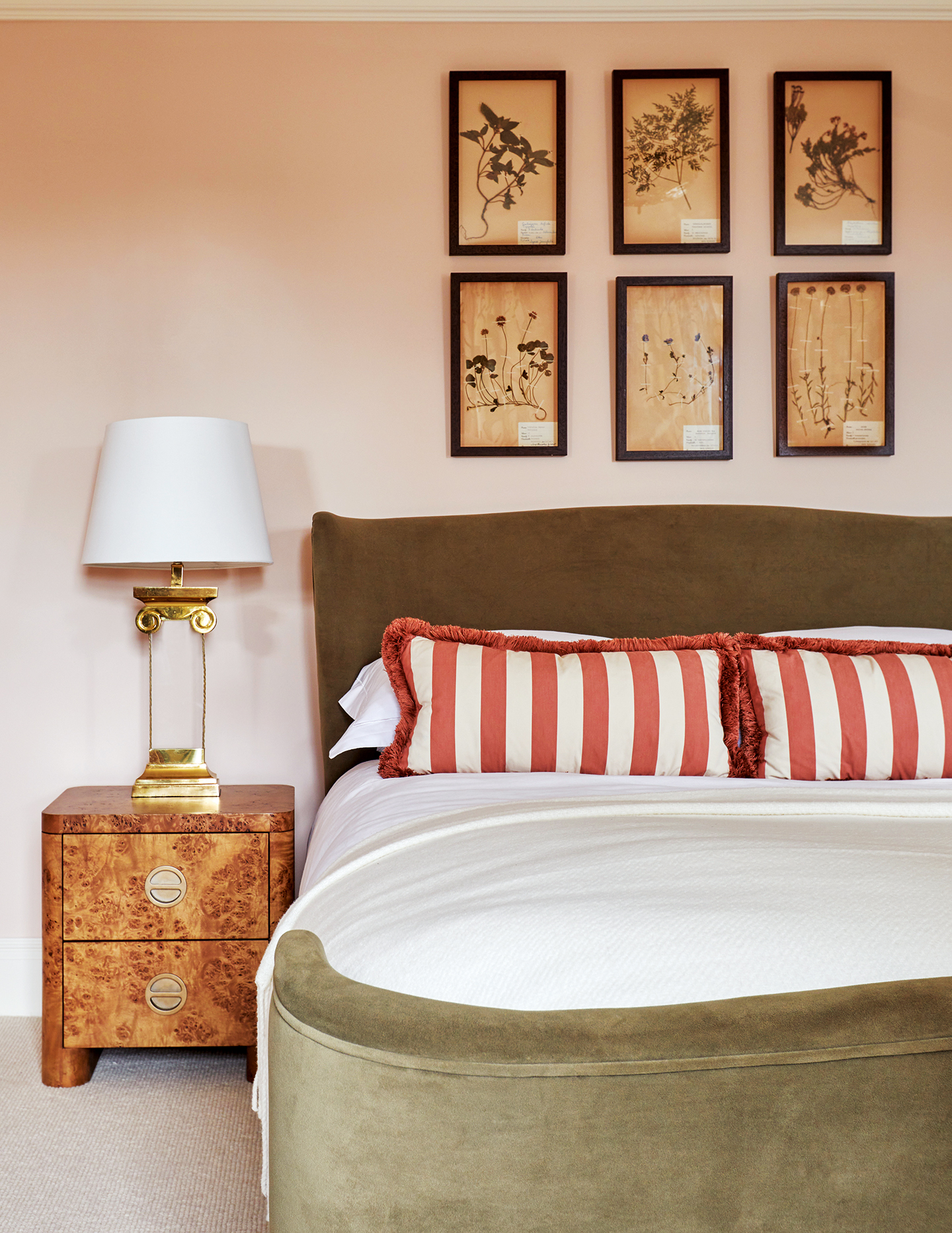 A pink bedroom with a double bed in green velvet, dressed with pink striped cushions with fringing. A series of six botanical prints hangs above. A burl wood bedside table is topped with a column-shaped glass table lamp