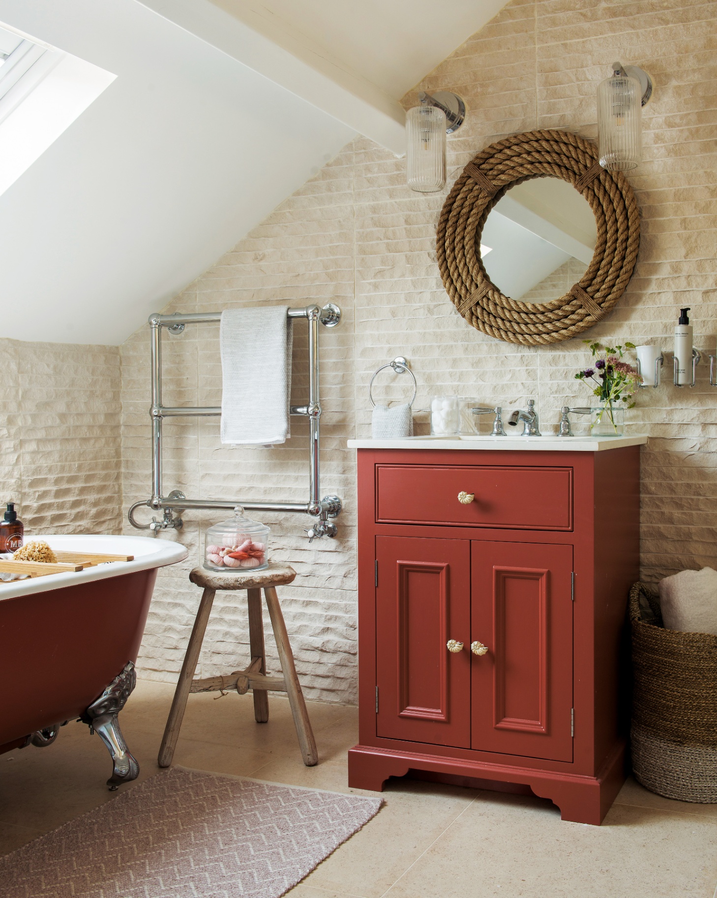 a bathroom with a red painted vanity unit with gold seashell door knobs, a large circle rope mirror and two glass ribbed wall lights above it and a red painted freestanding bath