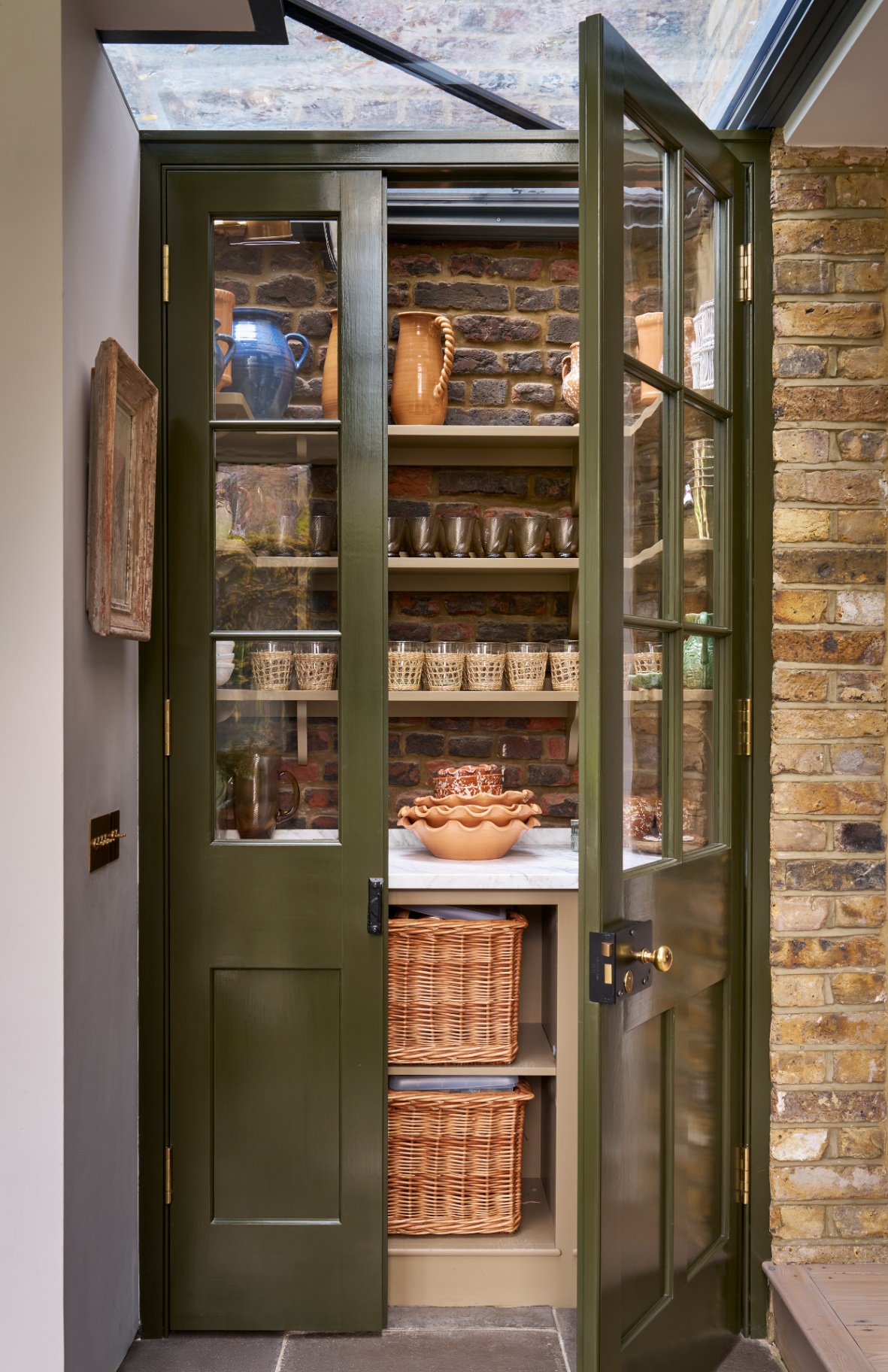 a pantry with dark green painted doors, 3 open shelves with glasses and jugs on, a white marble worktop with a stack of terracotta bowls and two wicker baskets underneath