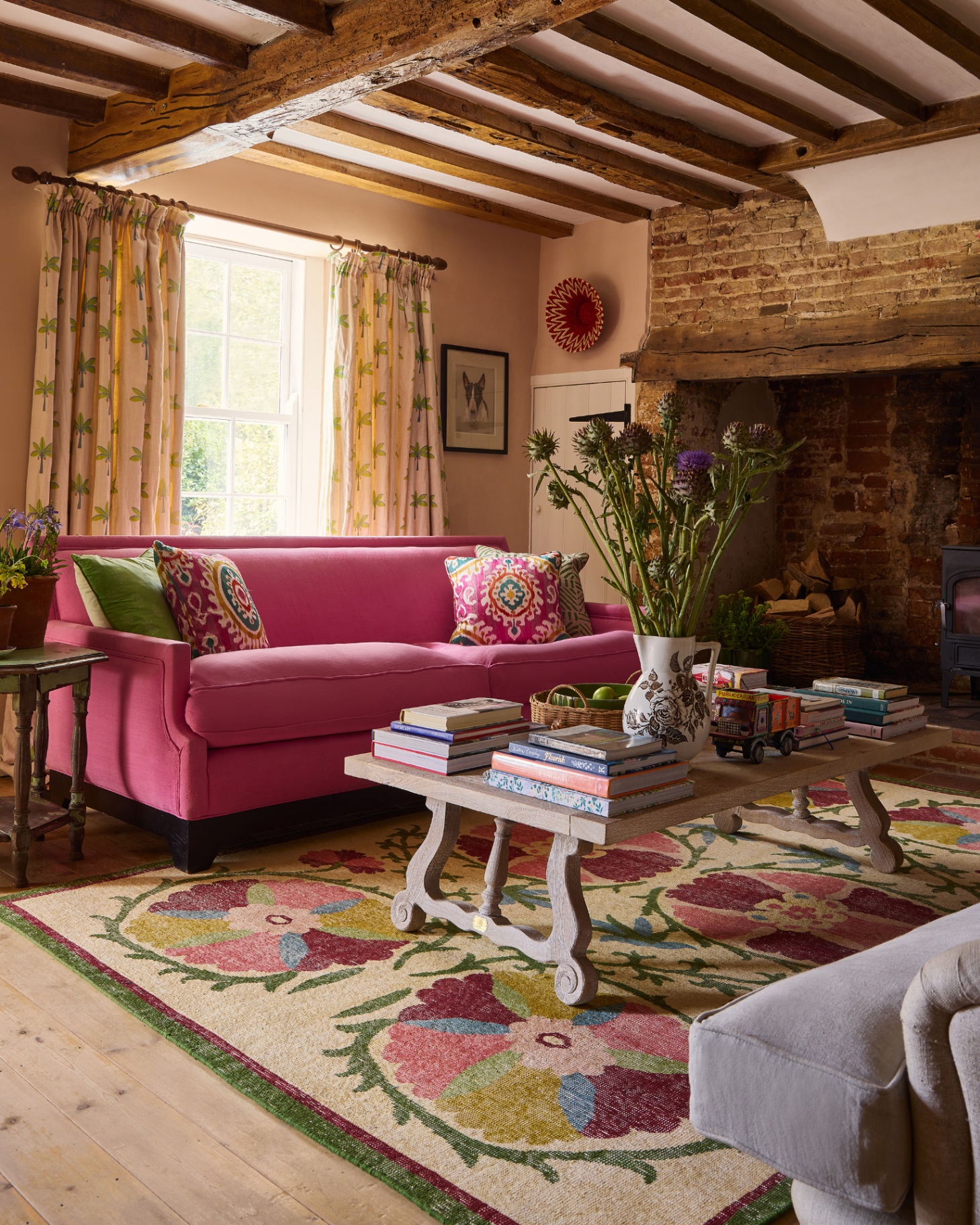 a living room with a bright pink sofa, a colourful floral patterned jute rug, a coffee table with piles of colourful books, a brick fireplace and wooden beams on the ceiling