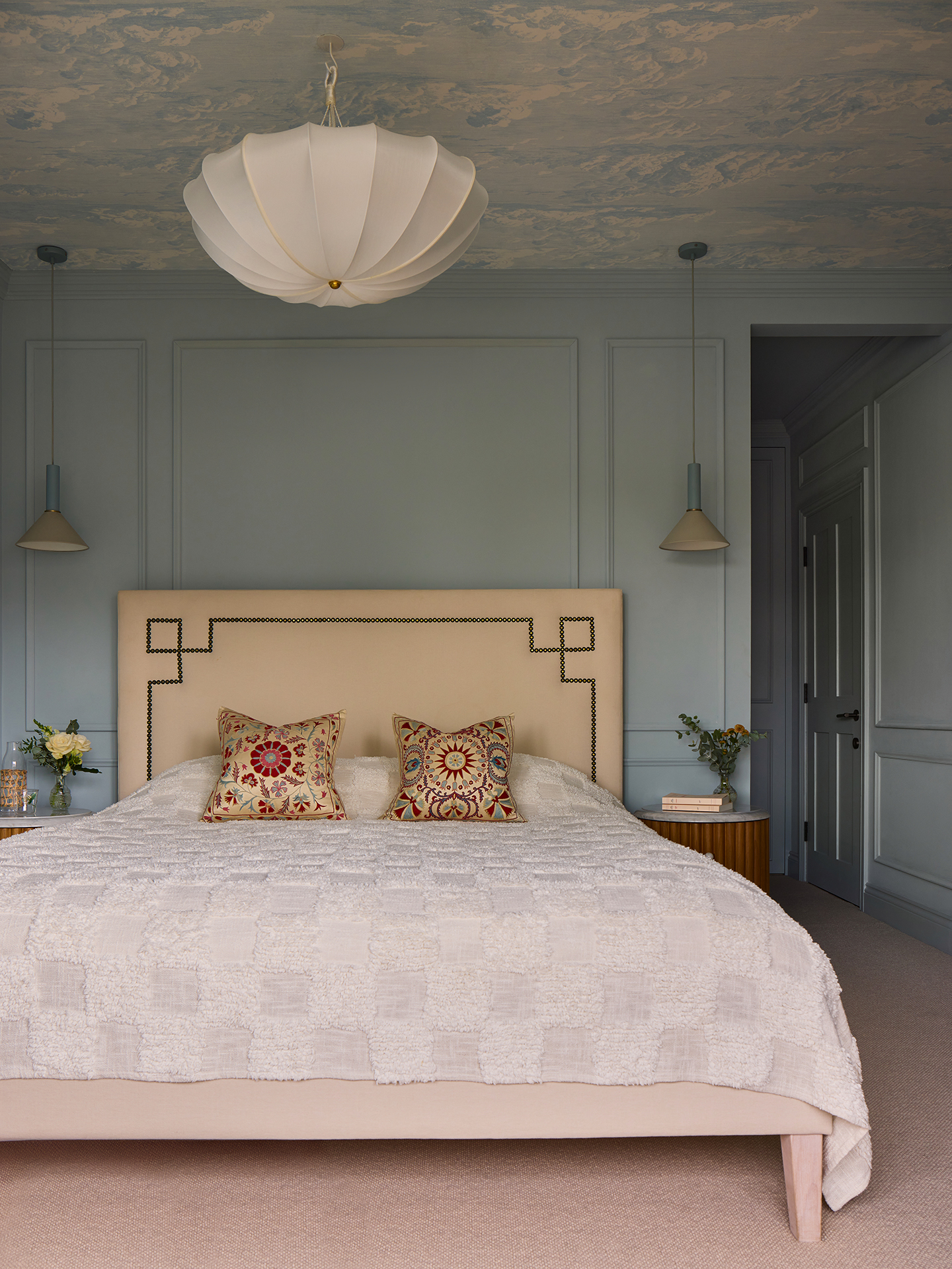 a large bedroom with a ceiling covered in cloud wallpaper. A large silk pendant light hangs in the middle of the room and two pendant lights hang either side of the double bed. The walls are painted in light blue and feature panelling details