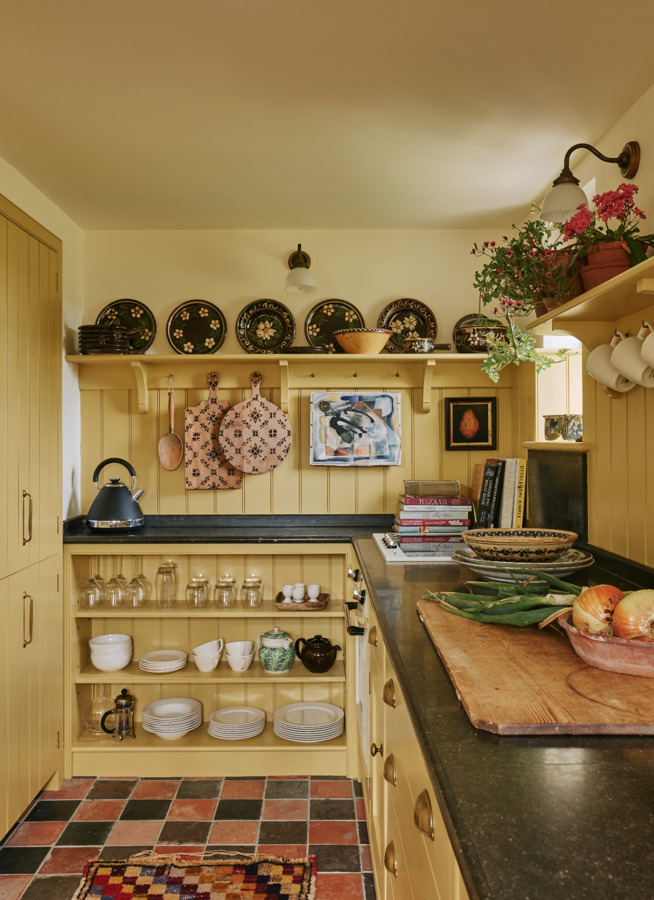 A small kitchen with pale yellow walls, cabinetry and ceiling, a black and terracotta checkerboard floor and decorative plates on the walls and shelves