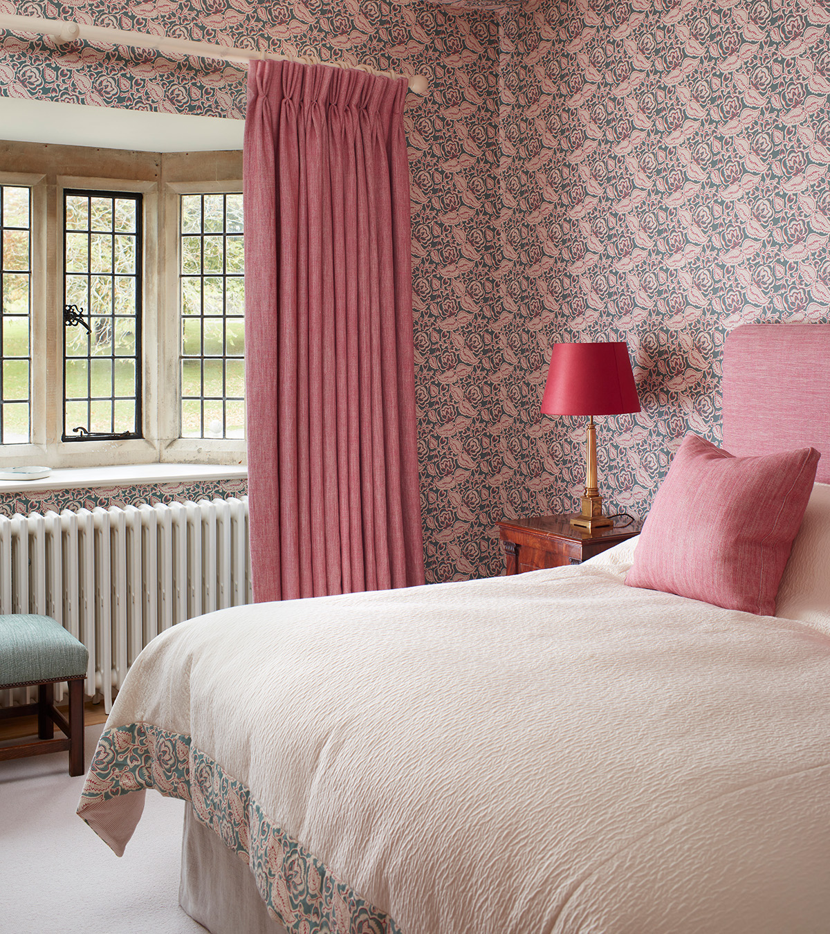a bedroom with pink curtains, pink cushions and a pink headboard, pink floral wallpaper and a double bed with white bed linen