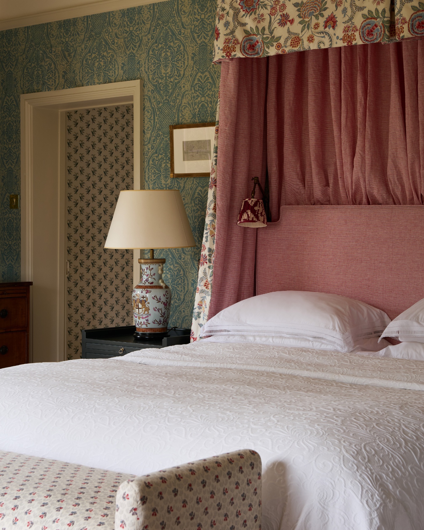 a bedroom with green botanical wallpaper, a bed with a large red and white striped headboard and canopy with a dark bedside table and a large ceramic patterned lamp