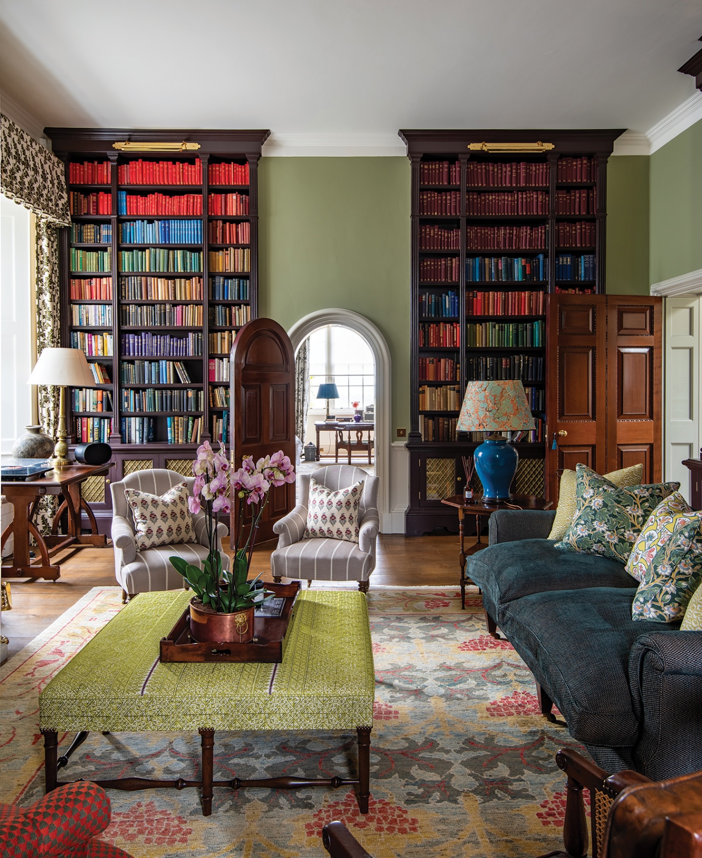 a large study with a seating area made up of a sofa, armchairs and upholstered ottoman anchored on a patterned rug in the foreground, and two large bookcases in the background either side of an open arched door
